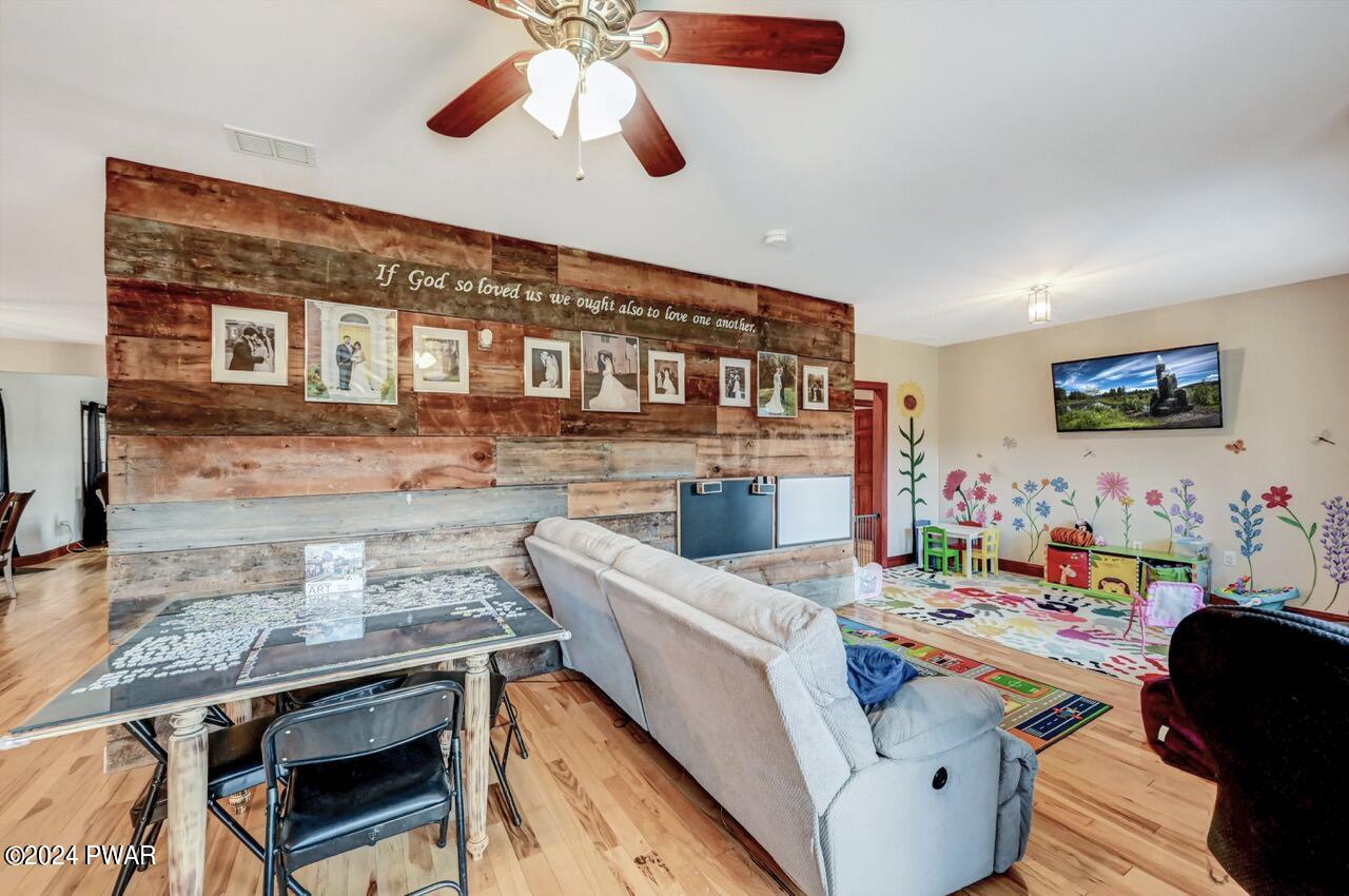 A living room with hardwood floors, a rustic wood accent wall displaying framed photos and a quote, a gray sofa, and a colorful children's play area with wall decals and a TV mounted on the right wall.