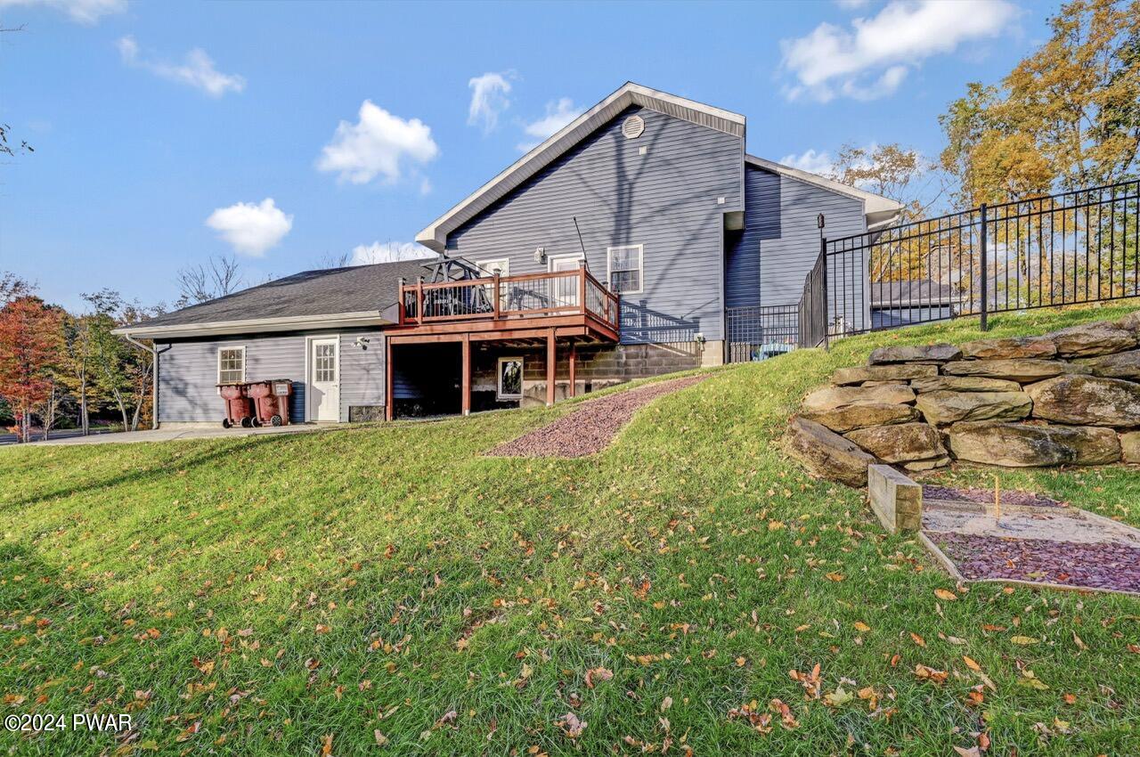 Exterior view of a gray house on a grassy slope with a wooden deck, stone retaining wall, and fenced area under a blue sky.