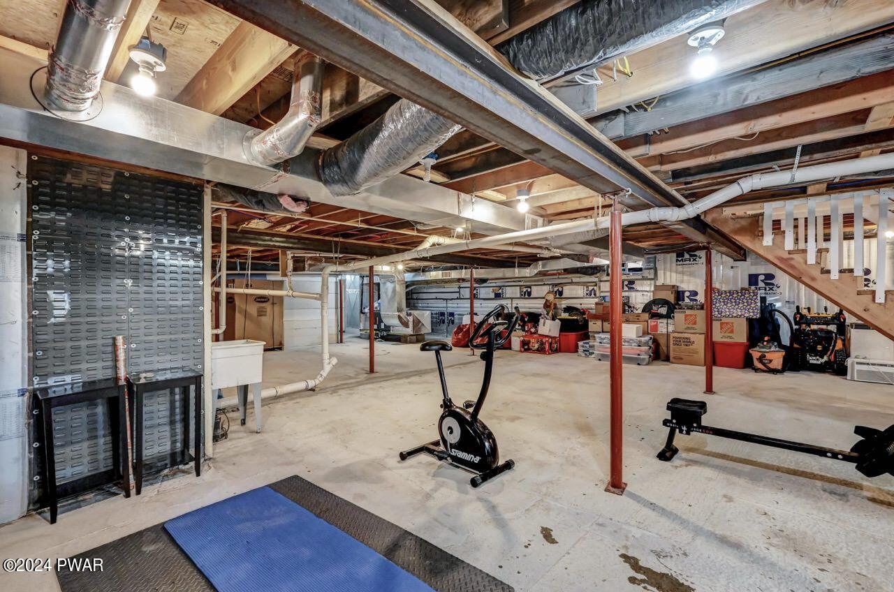Unfinished basement with exposed joists, ducts, support posts, concrete floor, storage boxes, exercise equipment, and stairs to upper level.