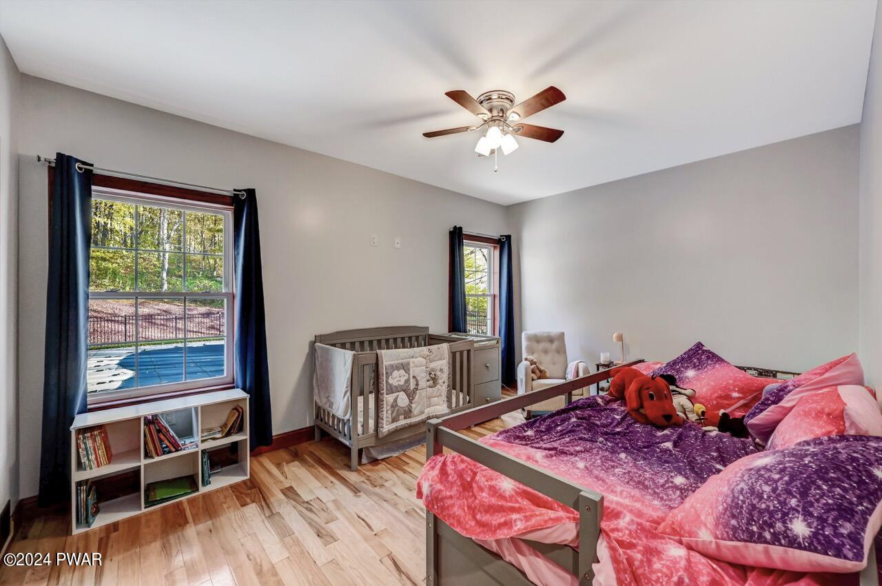 A child's bedroom with a crib, twin bed, bookshelf, two windows, and a ceiling fan.