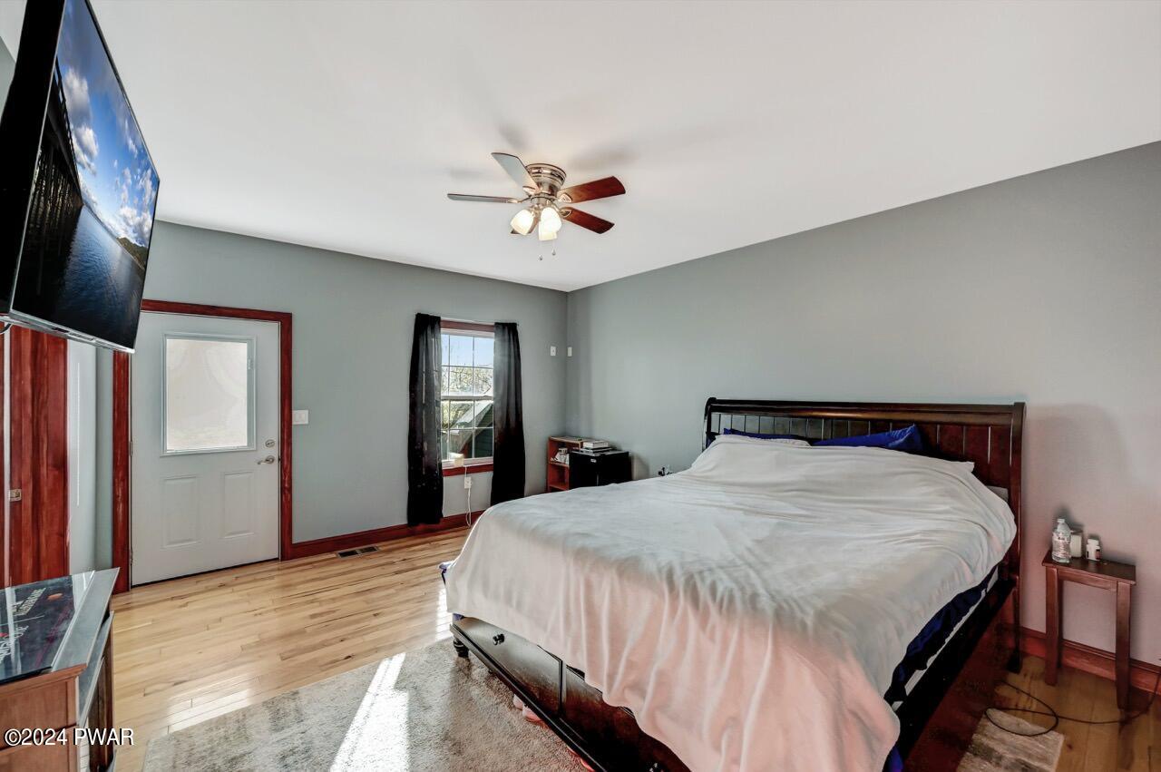 A furnished bedroom with a large bed, wall-mounted TV, gray walls, hardwood floors, and a ceiling fan.