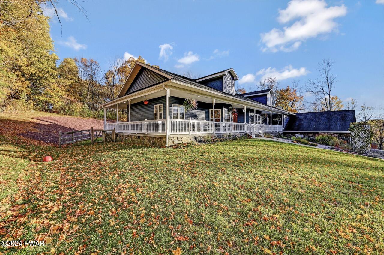 Exterior view of a dark-sided house with a wraparound white porch, sloped lawn, and autumn trees under a blue sky.