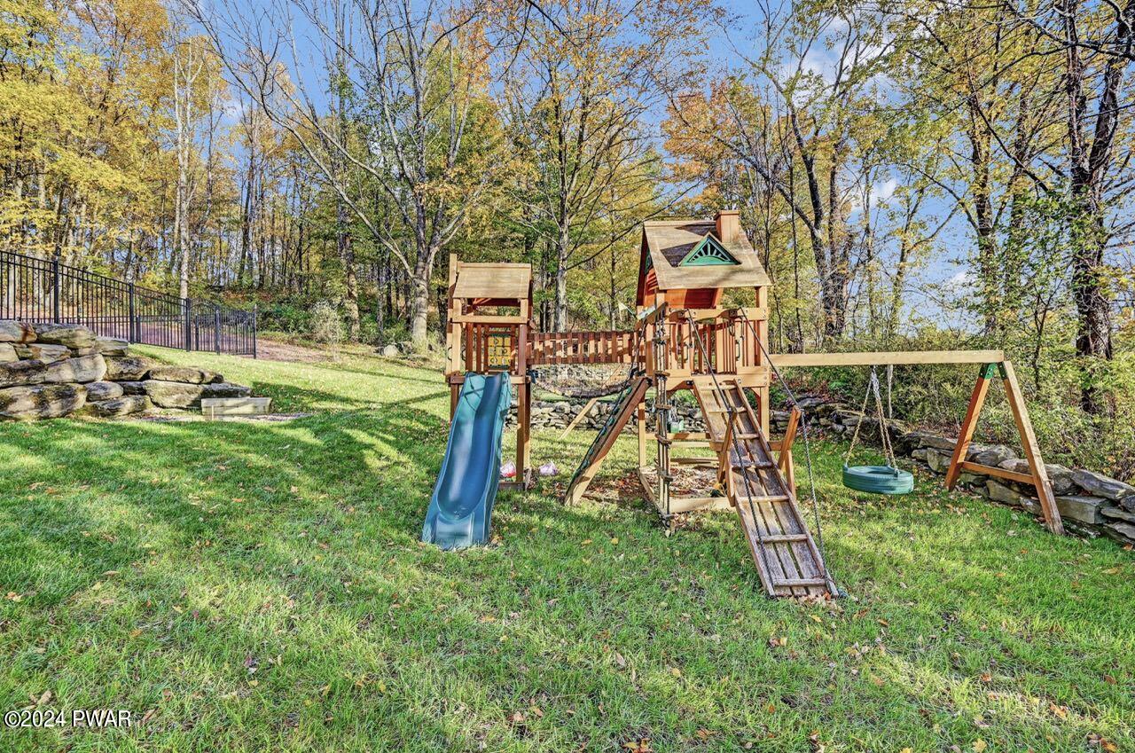 A backyard playground with a wooden playset, slide, ladder, and swings in a grassy yard surrounded by trees.