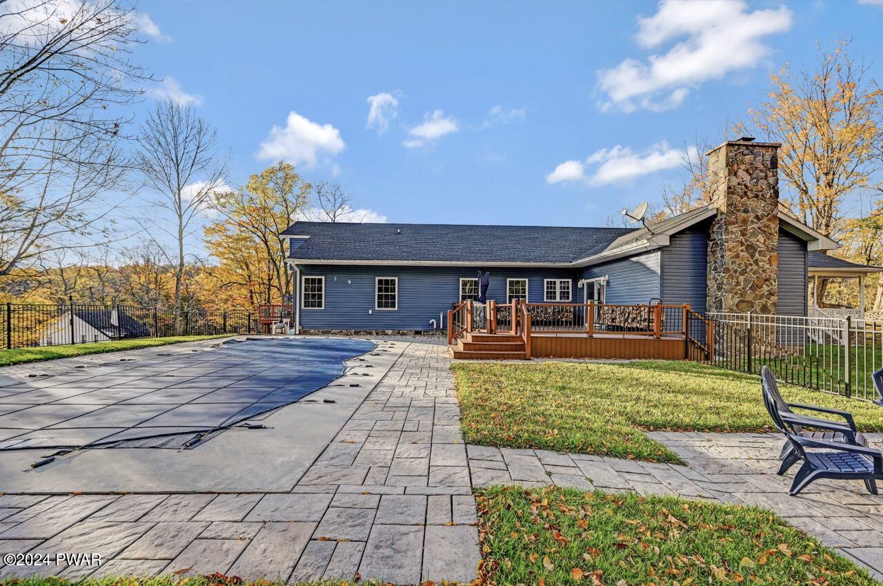 Rear exterior of a blue house with a stone chimney, deck, fenced yard, and paved patio area near a covered pool or tarp-covered surface.