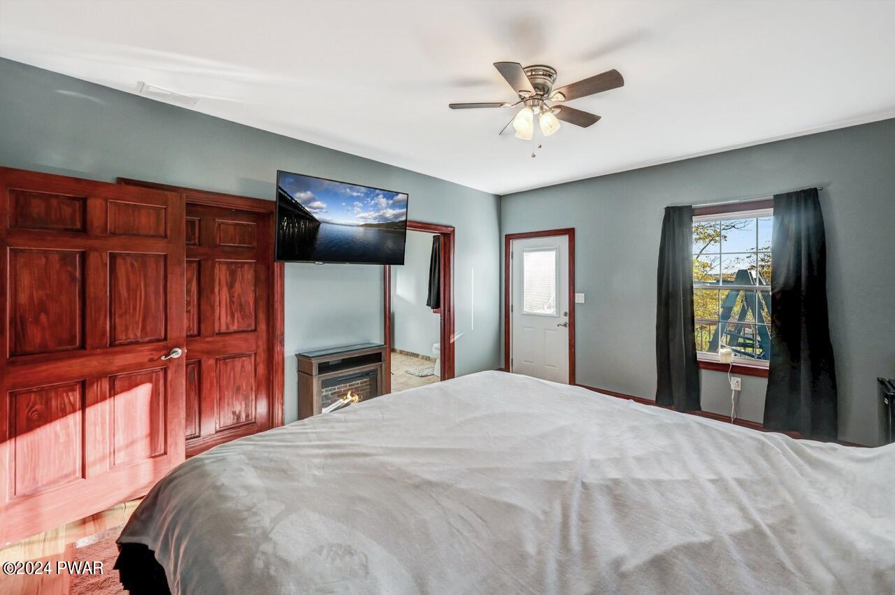 Bedroom with a bed in the foreground, wood closet doors, wall-mounted TV above an electric fireplace, a door to the hall or exterior, and a window with dark curtains.