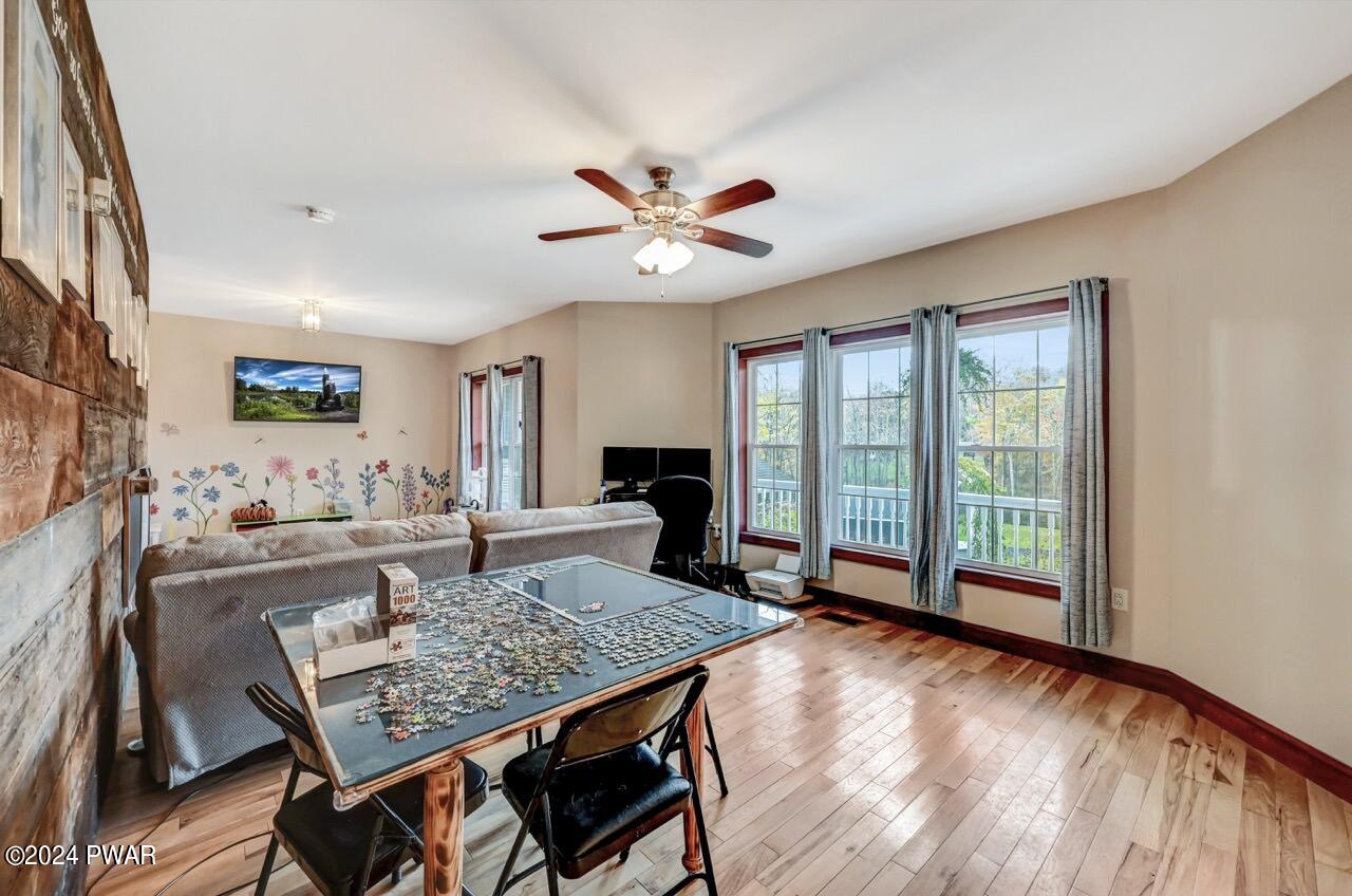 Wide-angle view of a living room with hardwood floors, a ceiling fan, large windows with curtains, a sofa, and a table set up with a jigsaw puzzle.