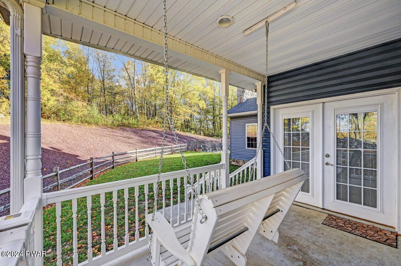 Covered porch with a hanging swing facing a grassy yard and wooded hillside, next to French doors.