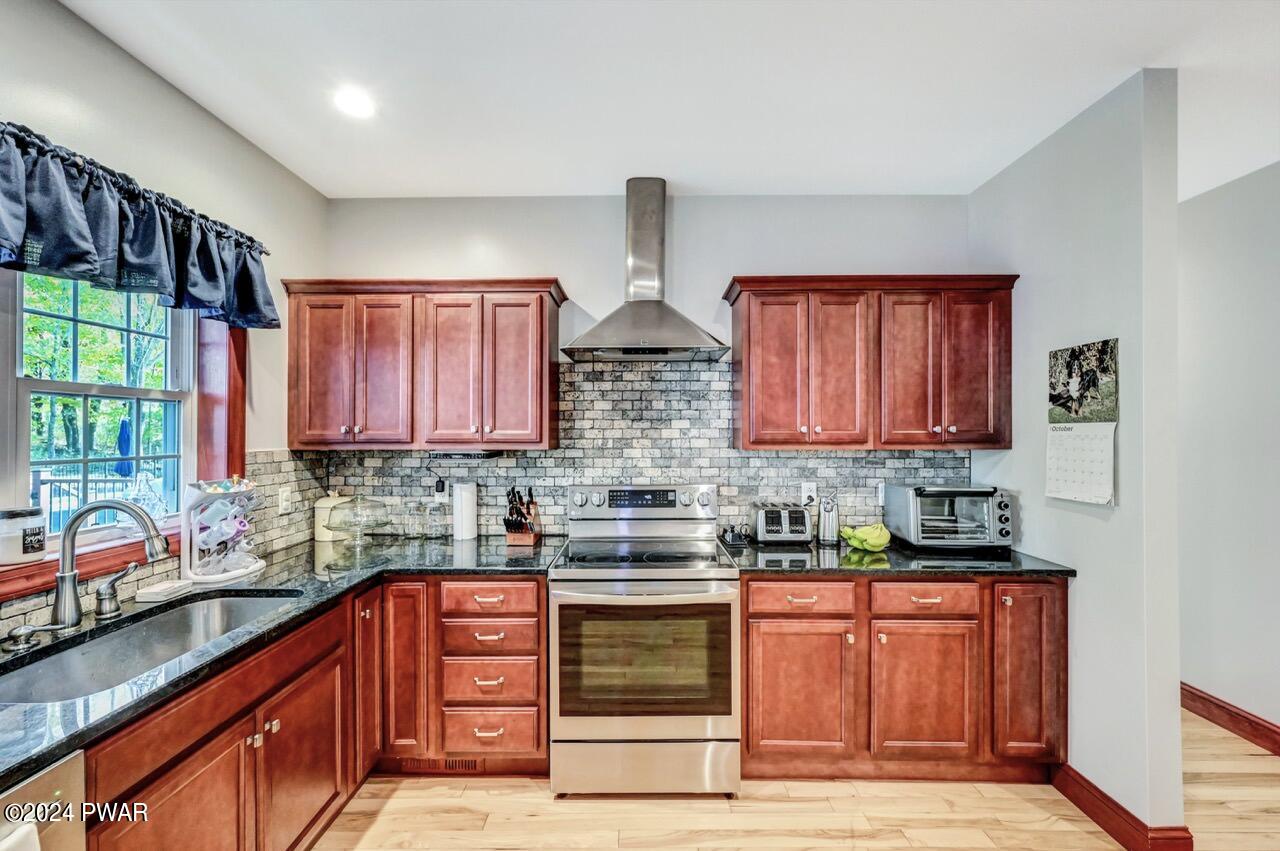 A bright kitchen with reddish wood cabinets, stainless steel range and hood, tiled backsplash, black countertops, and a sink under a window.