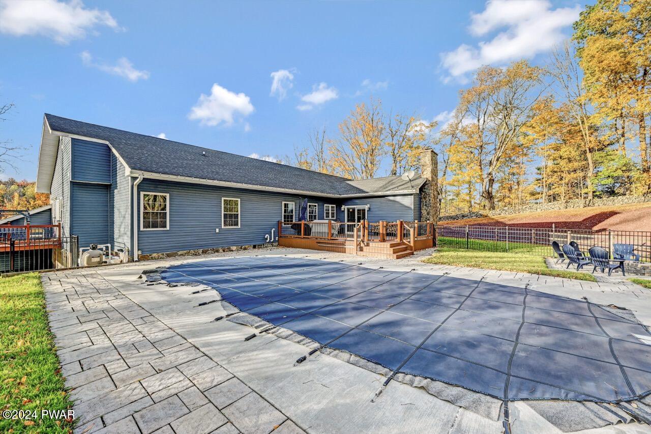 Back exterior of a blue house with a raised deck, stone chimney, paved patio, and a covered in-ground pool in the foreground.