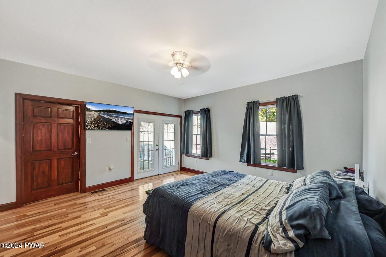 Furnished bedroom with hardwood floors, a bed, two windows with dark curtains, French doors, and a wall-mounted TV.