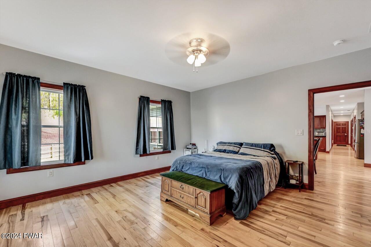 Spacious bedroom with hardwood floors, two windows with dark curtains, a bed against the far wall, and a doorway opening to a hallway and kitchen area.