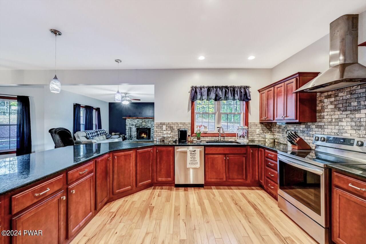 Kitchen with curved granite peninsula, cherry cabinets, stainless steel range and hood, tile backsplash, and view into a living room with a stone fireplace.