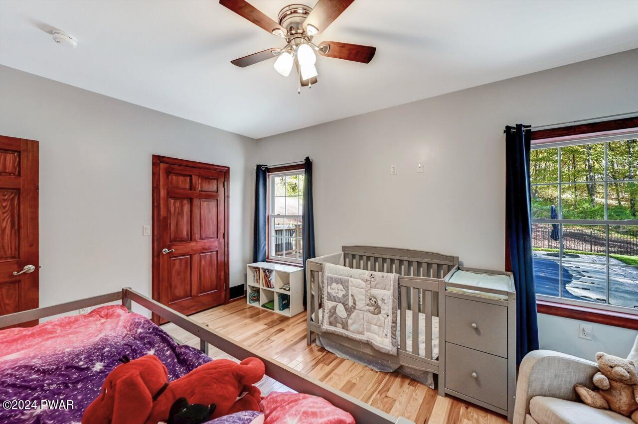 A bedroom with hardwood floors, gray walls, a ceiling fan, two windows with dark curtains, a crib, and a child’s bed with plush toys.