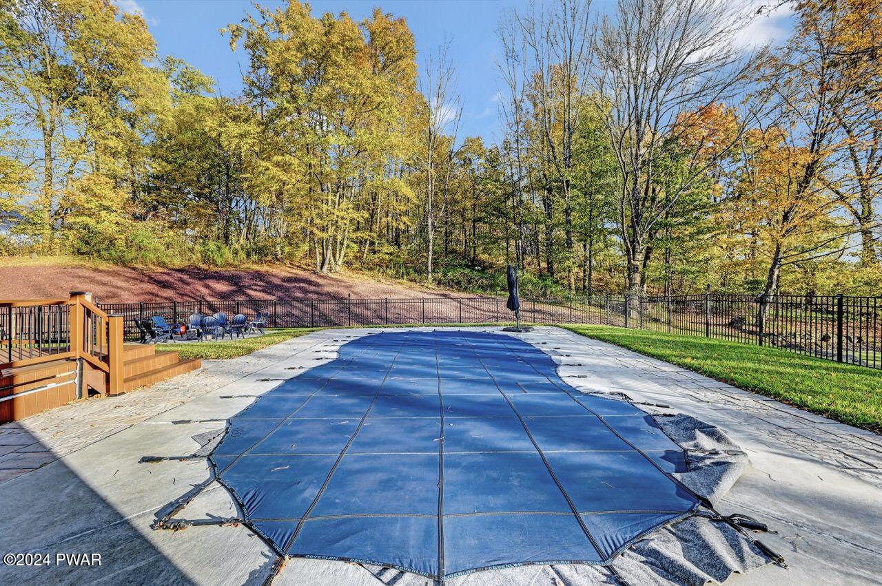Backyard patio with a covered in-ground pool, concrete decking, railing, and a fenced wooded backdrop in autumn.