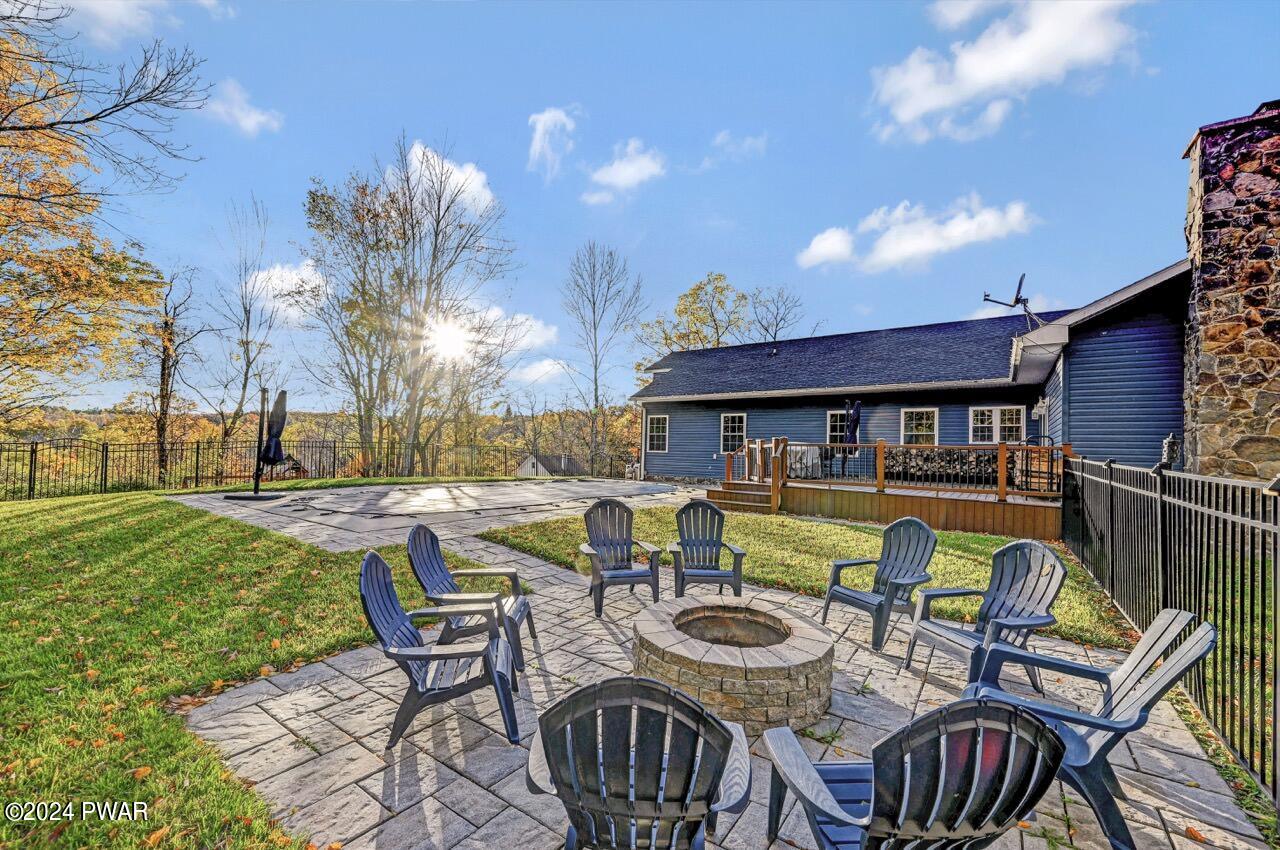 Stone patio with a circular fire pit surrounded by Adirondack chairs beside a blue house and deck, with a grassy yard and trees in the background.