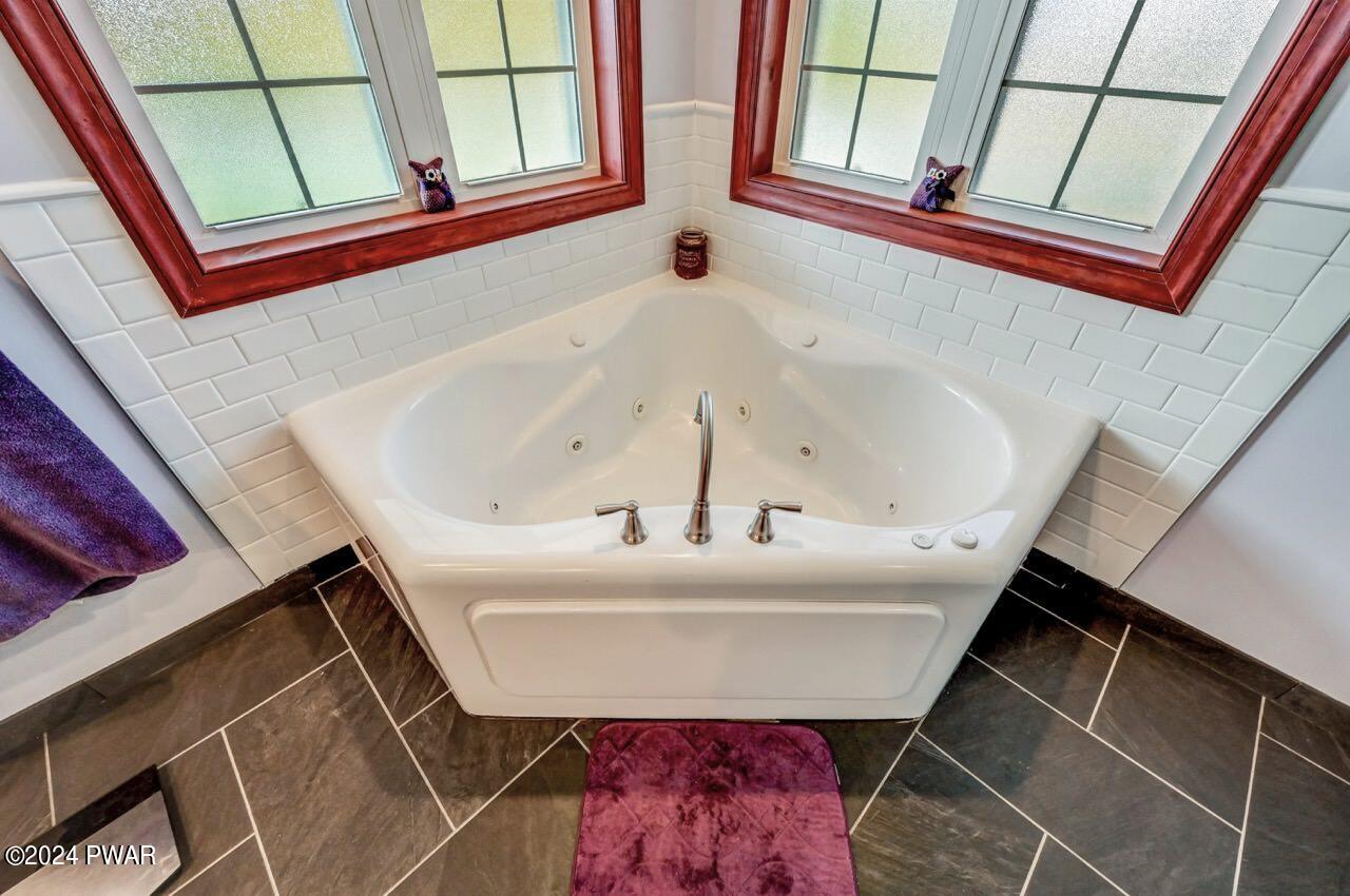 Corner bathroom with a white jetted soaking tub below two frosted windows, tiled walls, and dark floor tile.