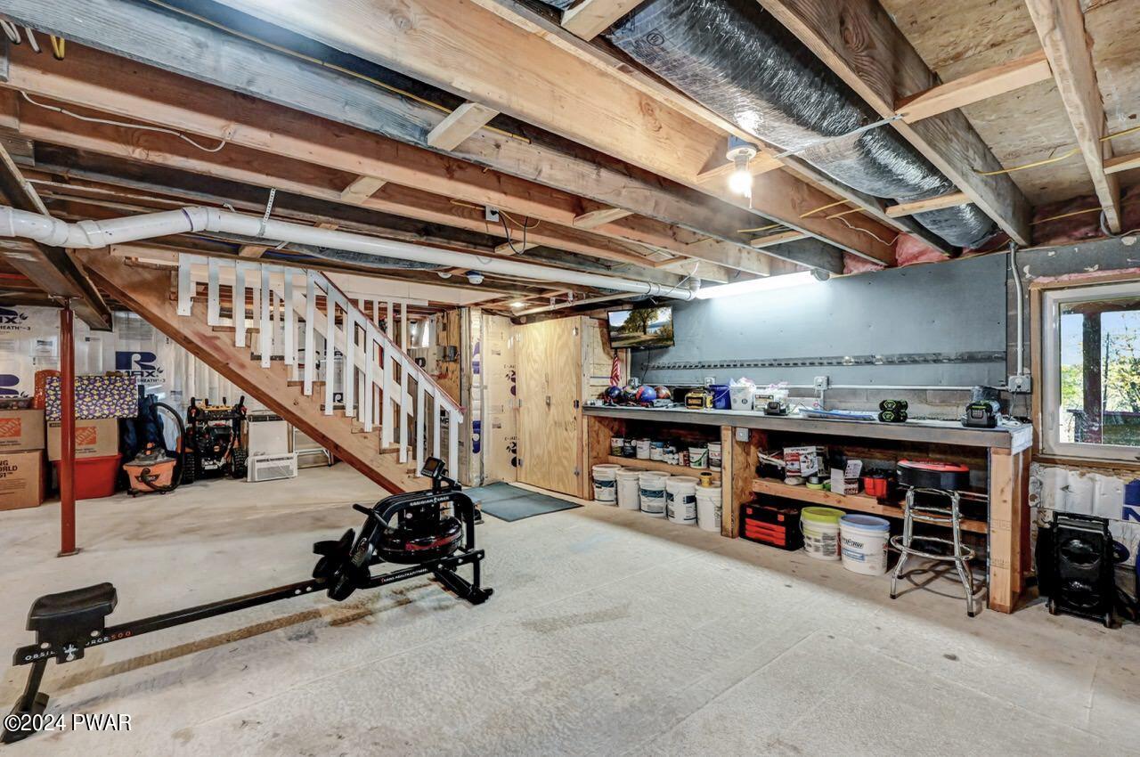 Unfinished basement with exposed ceiling joists, staircase, workbench, storage shelves, and exercise equipment.