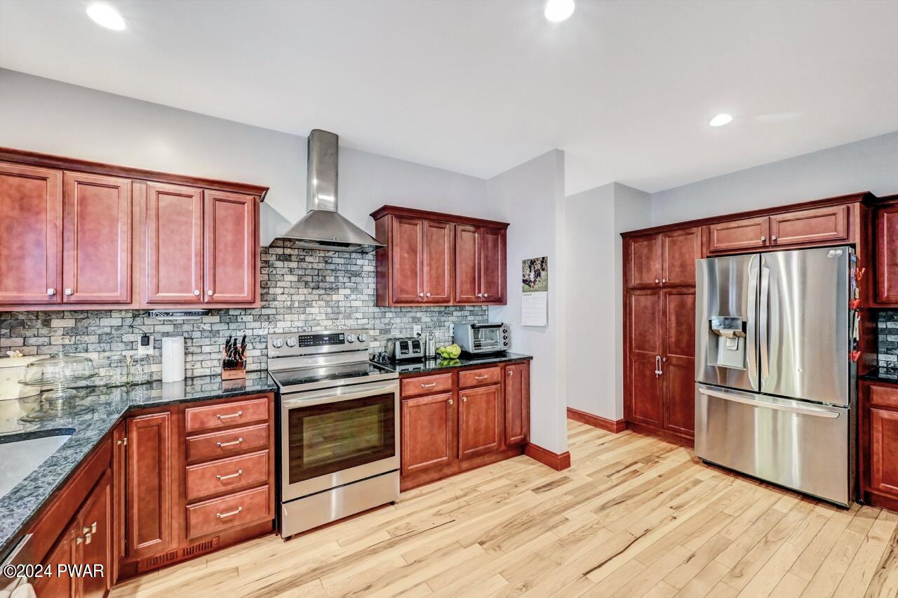 Bright kitchen with reddish wood cabinets, stainless steel refrigerator and range, stone backsplash, and hardwood floors.