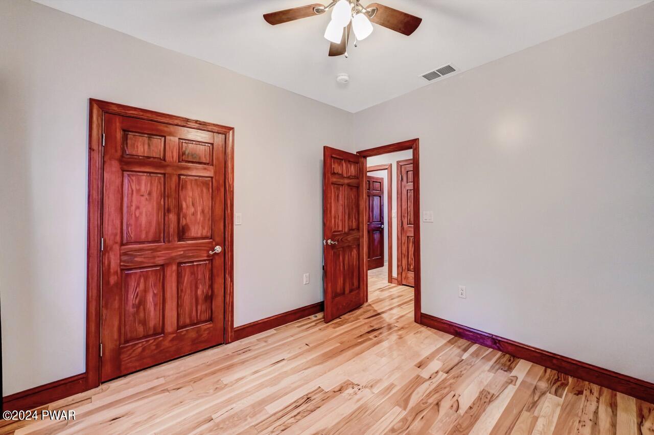 Empty bedroom with light gray walls, hardwood floors, wood trim, ceiling fan, and two wooden doors, one open to a hallway or adjoining room.