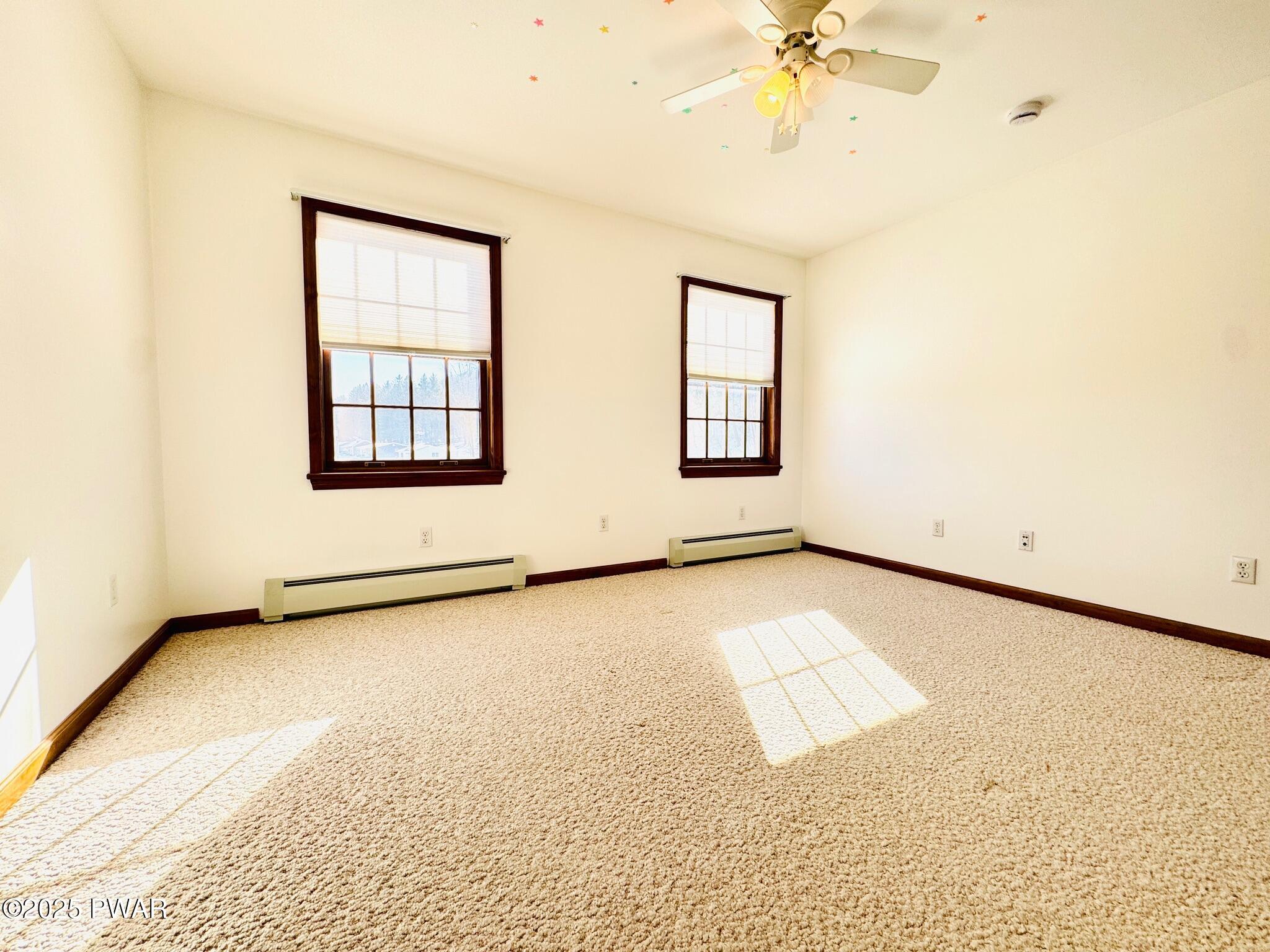 Empty carpeted bedroom with two windows, ceiling fan, and baseboard heaters.