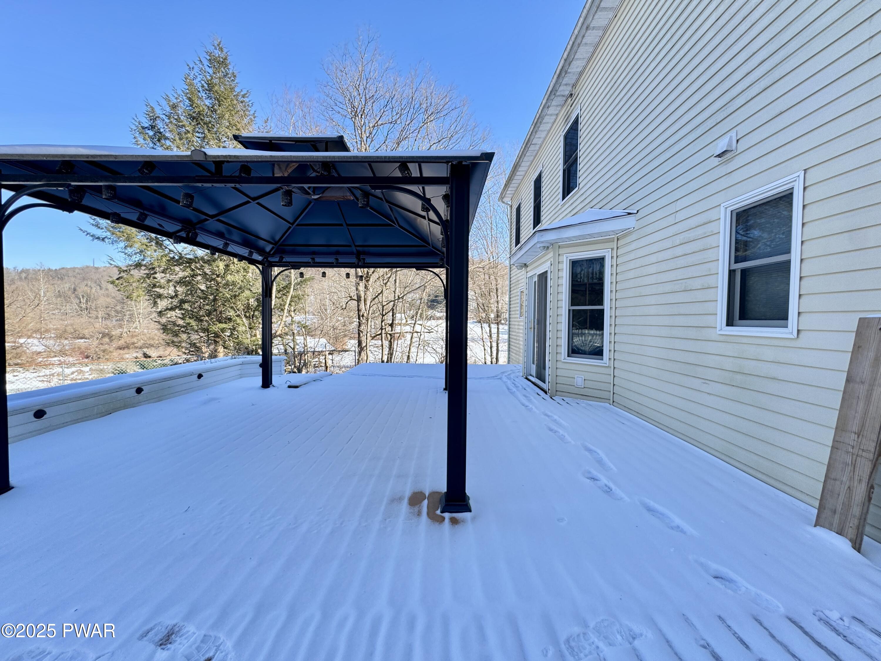 Snow-covered outdoor deck beside the rear exterior of a house, with a dark metal gazebo or pergola structure and sliding glass door.