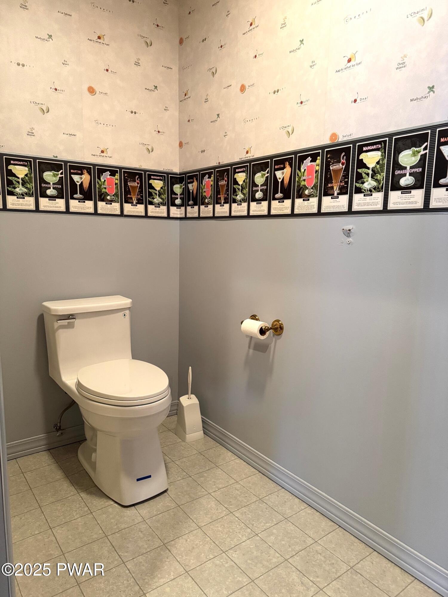 A small bathroom with a white toilet, gray walls, tile floor, and decorative cocktail-themed wallpaper border near the ceiling.