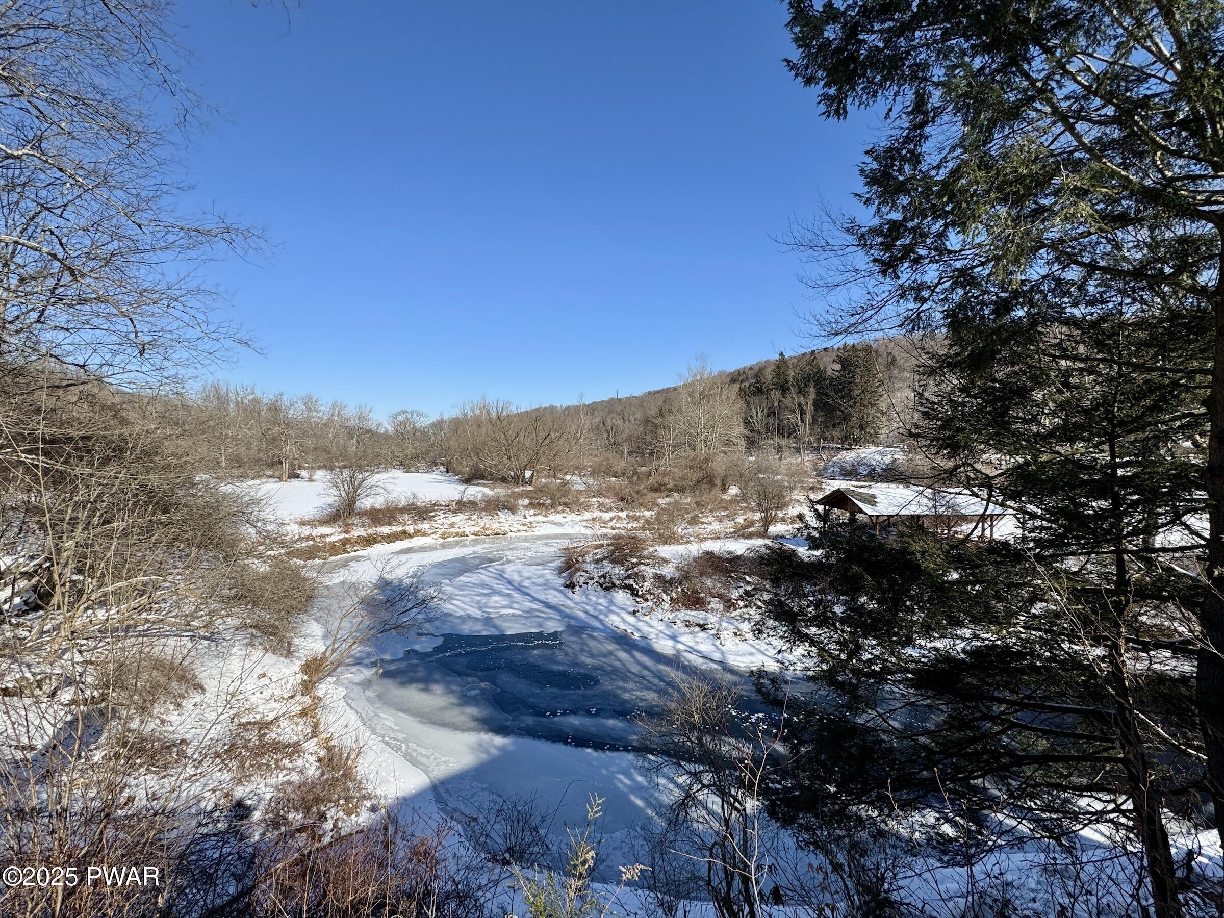 Snow-covered landscape with a partially frozen pond, leafless trees, evergreen branches framing the view, and a small roofed structure on the right under a clear blue sky.