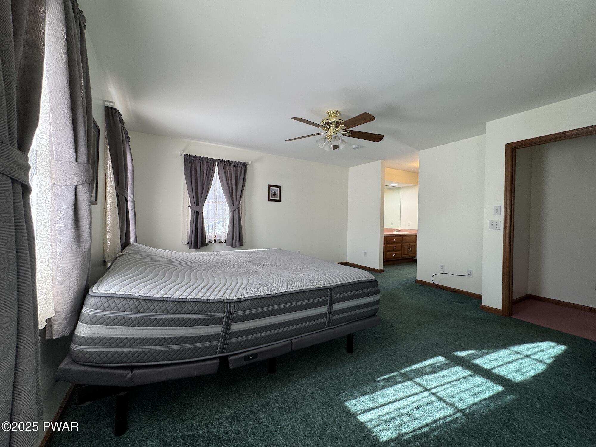 A bedroom with a bed, ceiling fan, green carpet, curtained windows, and an open doorway to a bathroom vanity area.