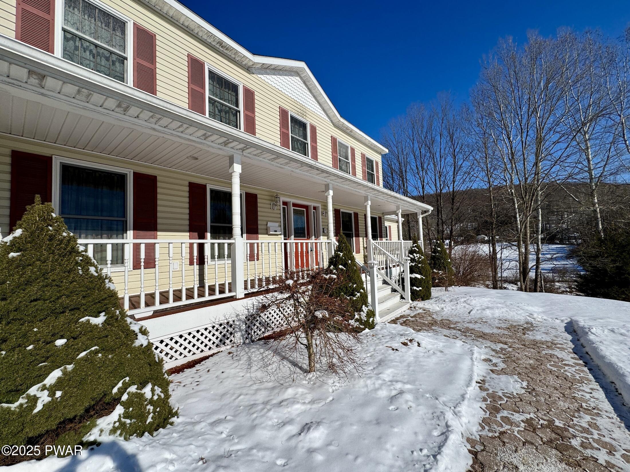 Two-story house exterior with a covered front porch, red shutters, snow-covered yard, and a stone walkway under a bright blue sky.