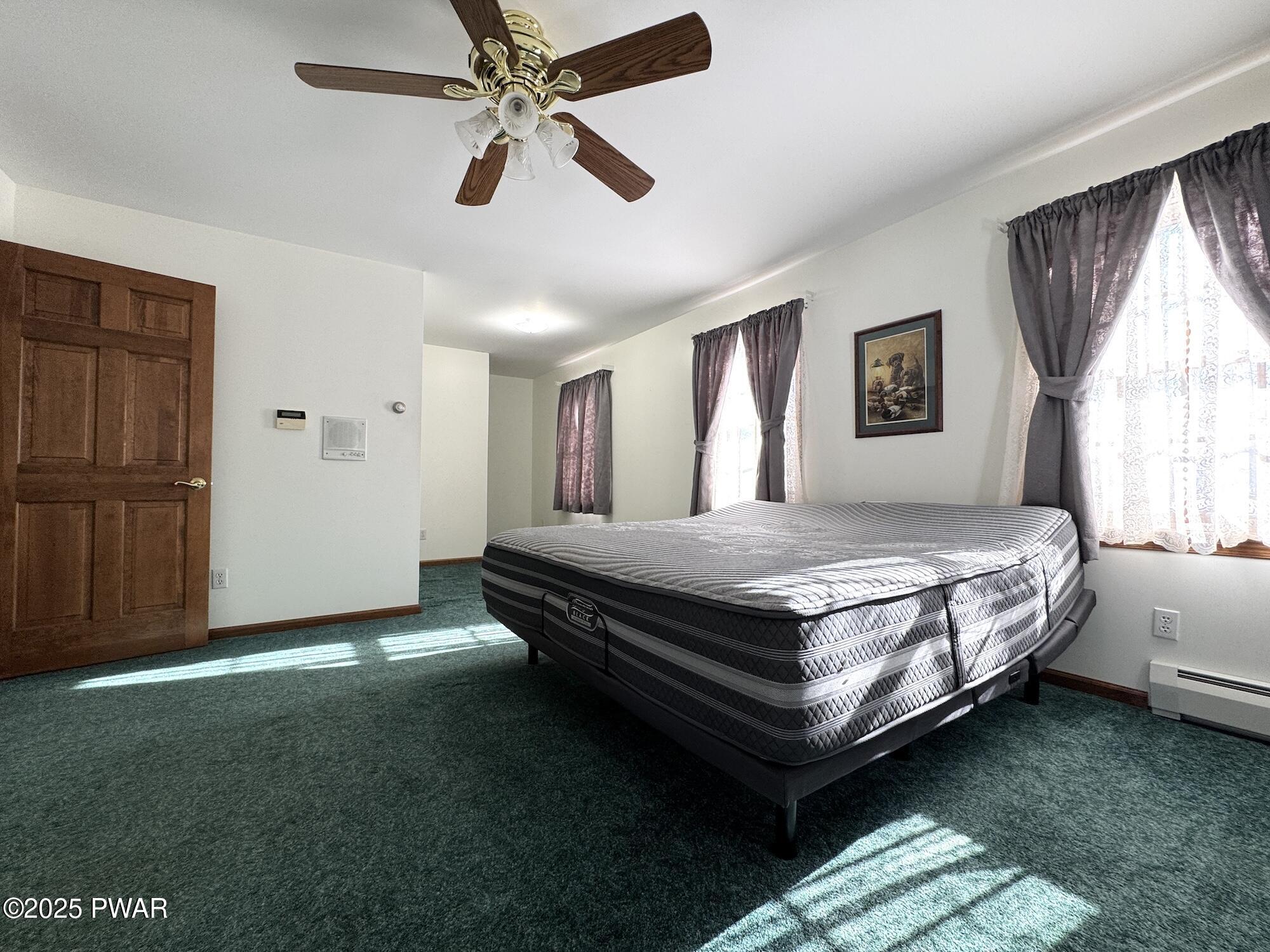 A bedroom with a bed, green carpet, white walls, curtained windows, a ceiling fan, and a wood door.