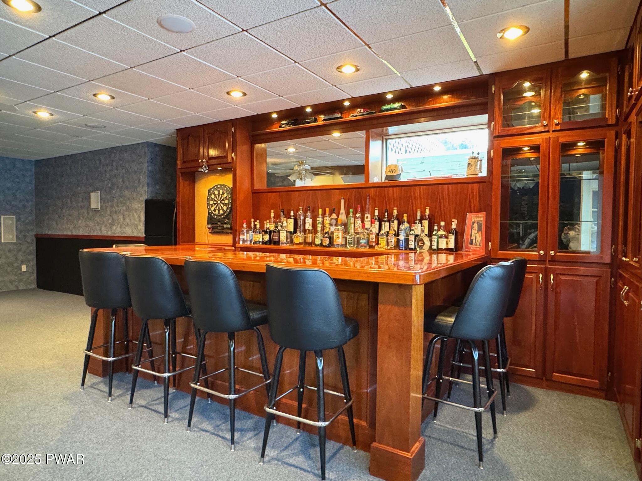 Finished basement bar with wood cabinetry, a polished bar counter, bottle display, and several barstools.
