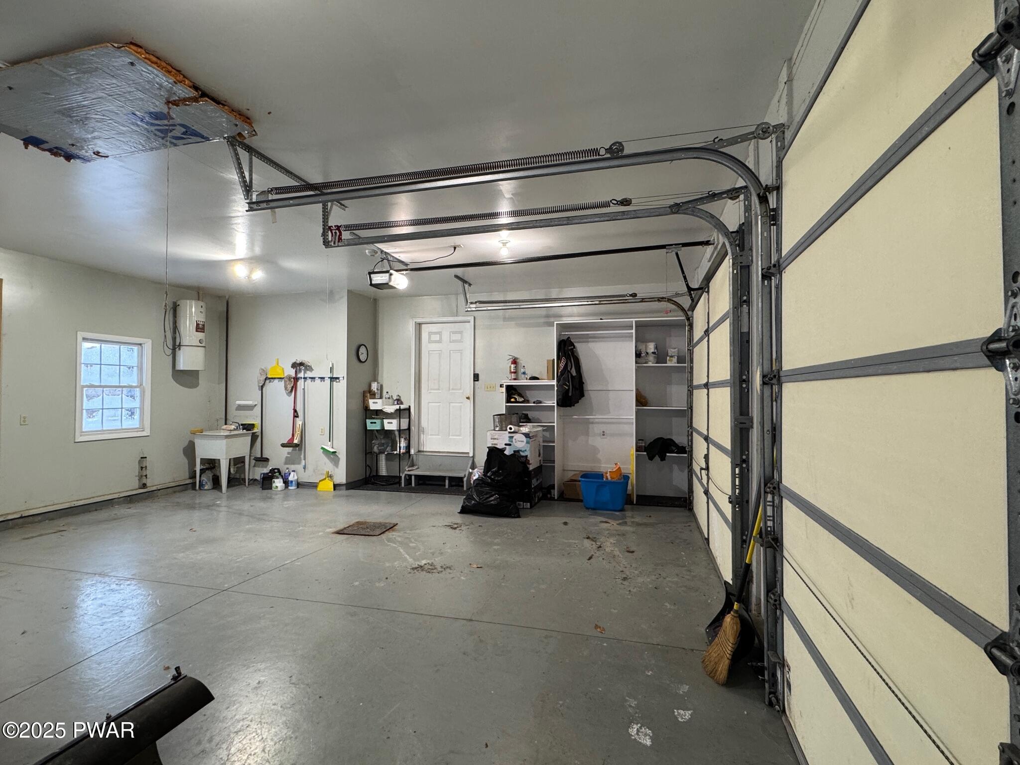 Interior view of a garage with a concrete floor, open garage doors, storage shelves, a side window, and utility items along the walls.