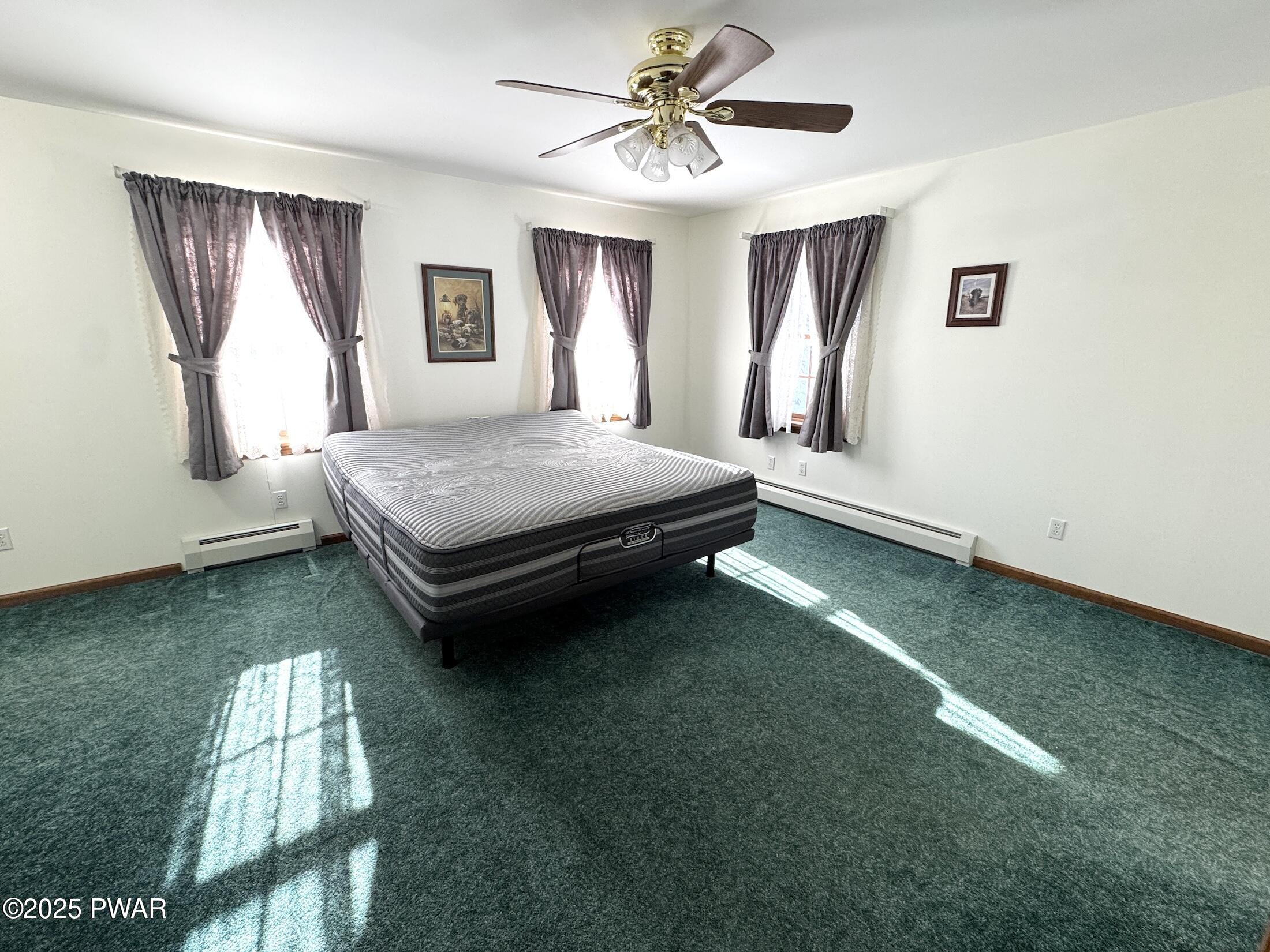 An empty bedroom with a bed, teal carpet, three windows with curtains, and a ceiling fan.