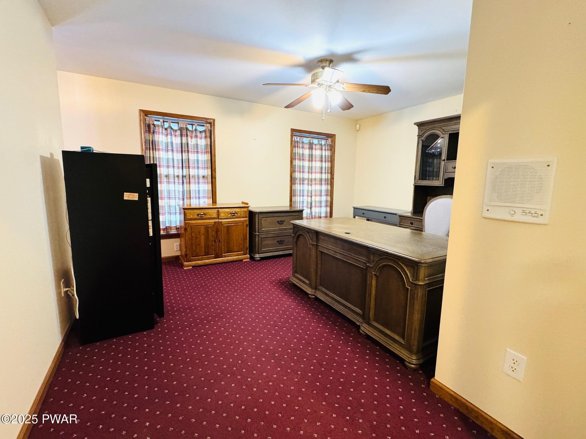 A furnished office with burgundy carpet, cream walls, two windows, a ceiling fan, and several wooden desks and cabinets.