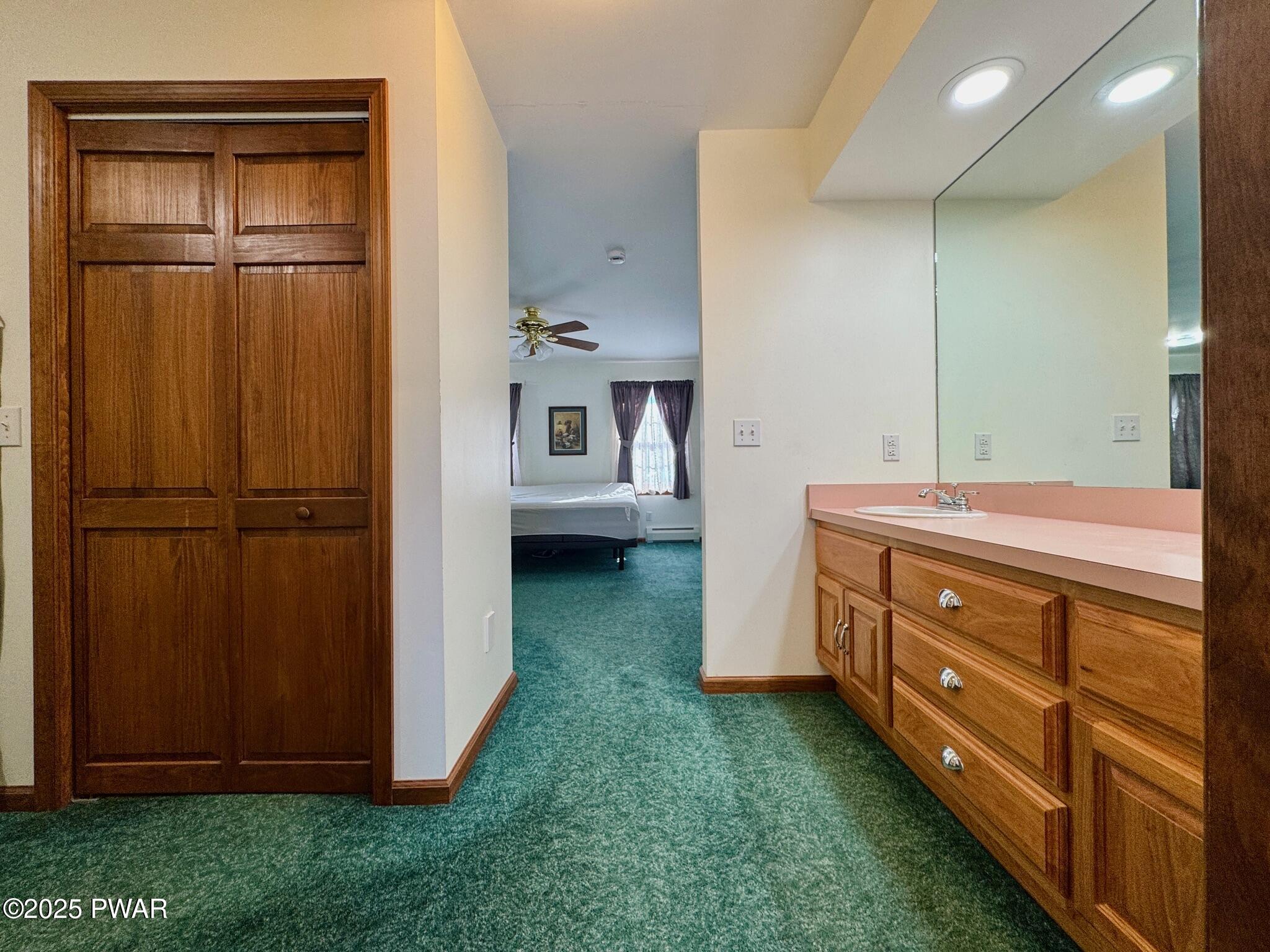 Bathroom with wood vanity, pink countertop, large mirror, green carpet, and a view into a bedroom beyond.