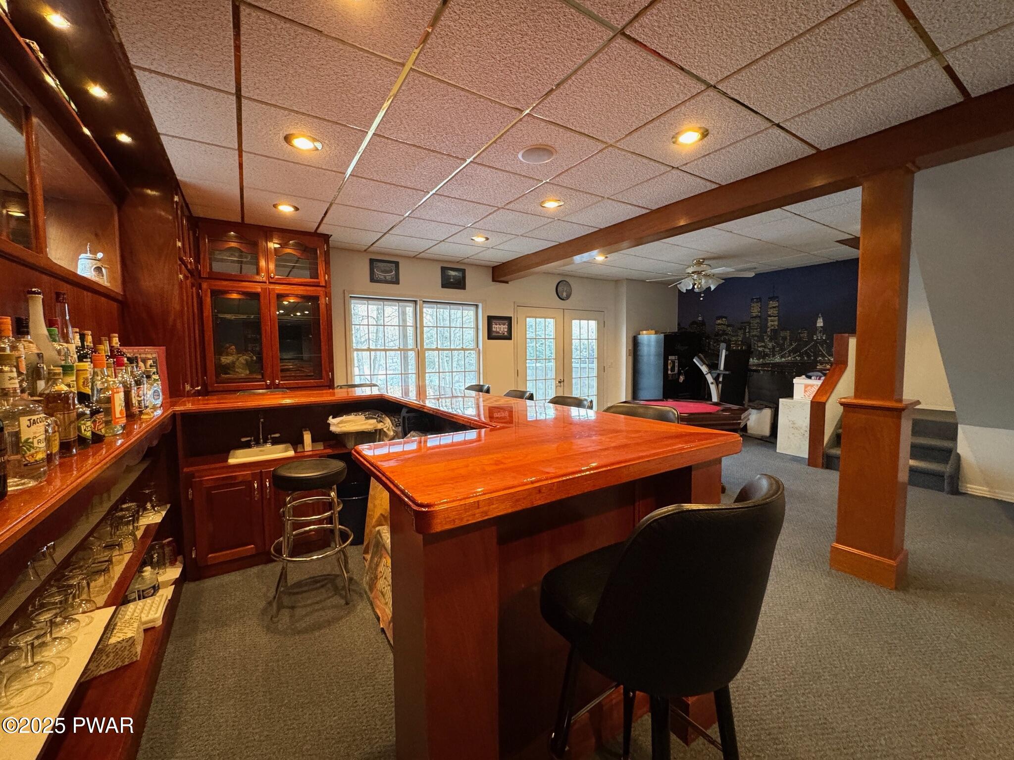 Finished basement bar area with wood bar, shelves of liquor bottles, bar stools, carpeted floor, and a wall mural.