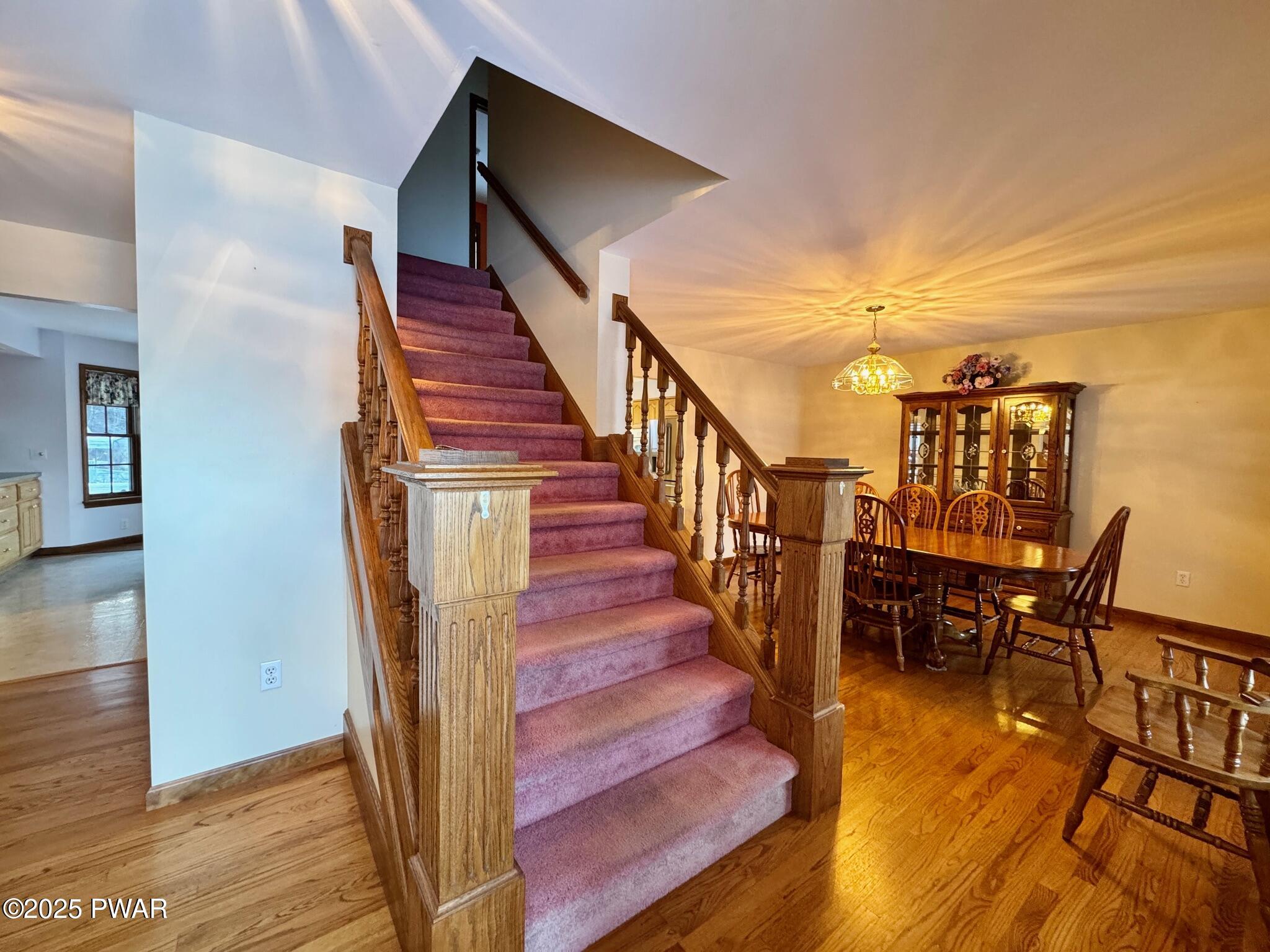 Dining room with wood floors, staircase with pink carpet runner, chandelier, and china cabinet.
