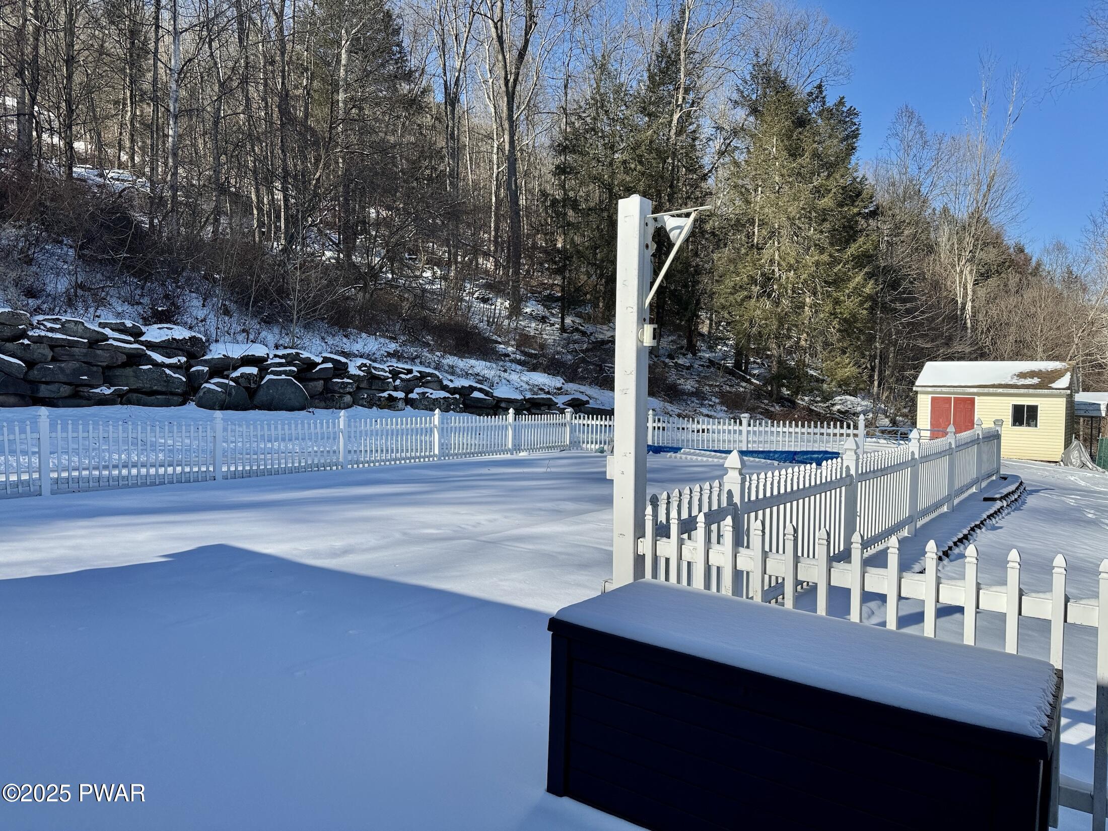 Snow-covered fenced backyard with a pool area, white picket fencing, a small shed, and wooded hillside in the background.