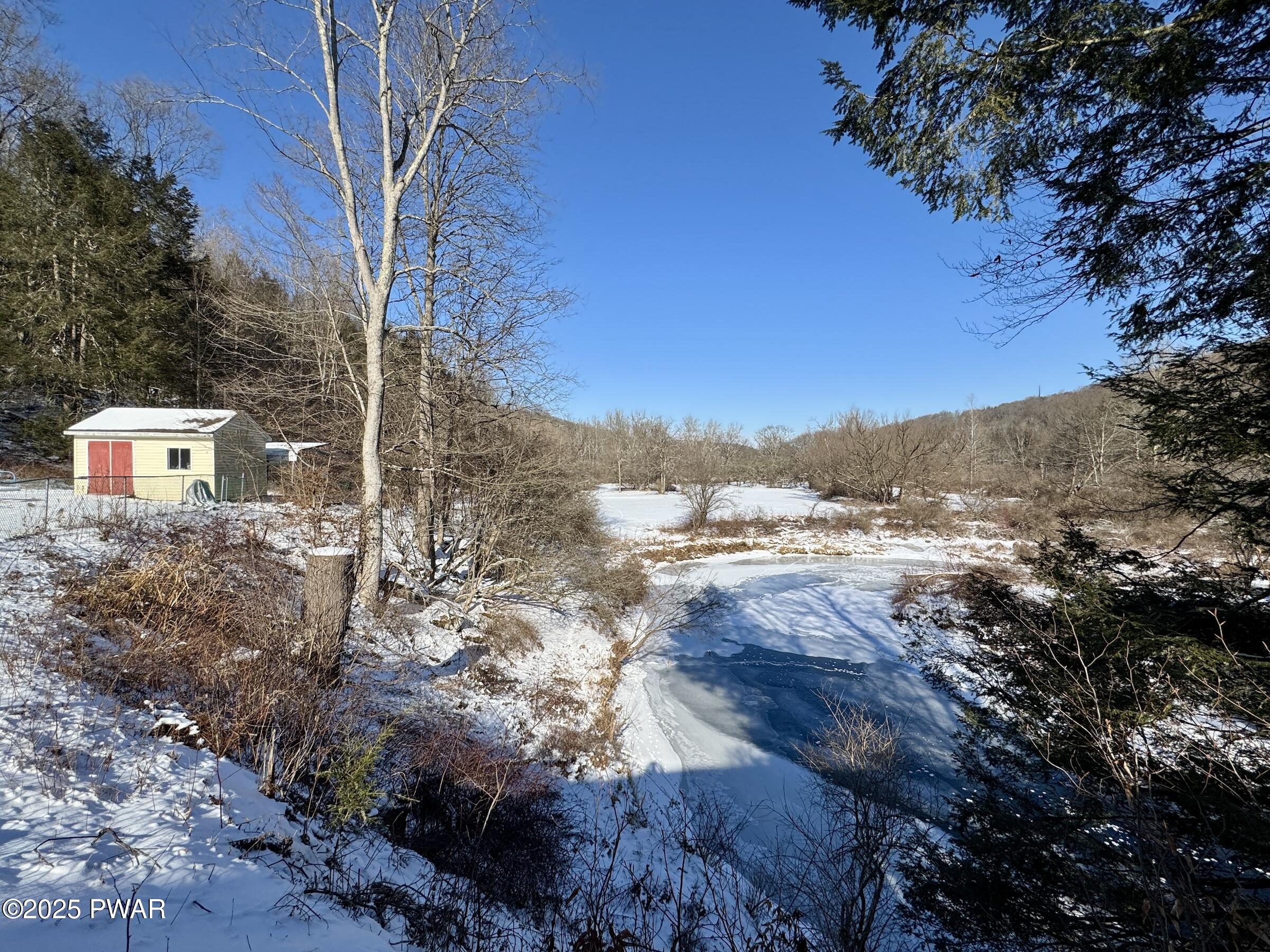 Snow-covered rural landscape with a frozen pond or creek, trees, and a small shed on the left under a clear blue sky.