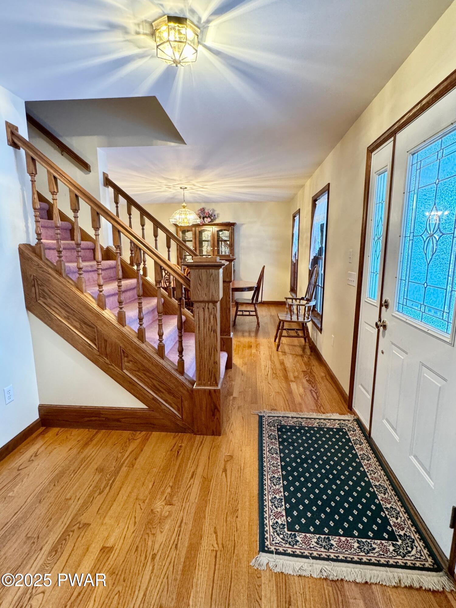 A home entry foyer with hardwood floors, a staircase with pink carpeted steps and wood railing, a front door with decorative glass, and a small table and chairs in the hall.