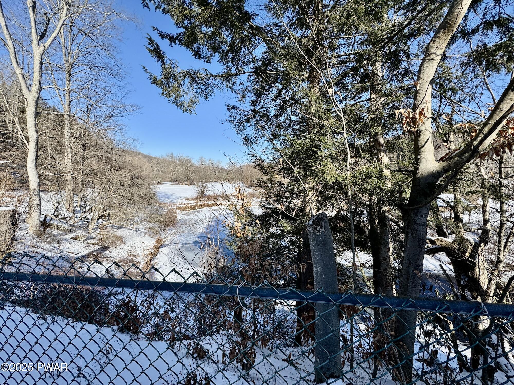 Snow-covered wooded lot with trees, a creek or wetland in the distance, and a chain-link fence in the foreground.