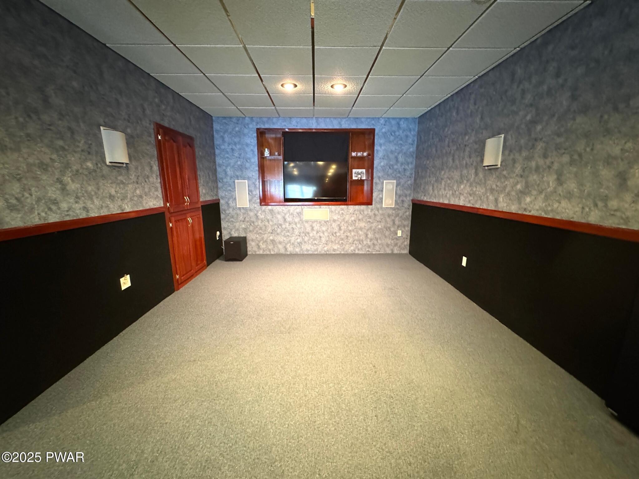 A carpeted home theater room with a large wall-mounted TV, built-in wood trim, surround speakers, and dark wainscoting.