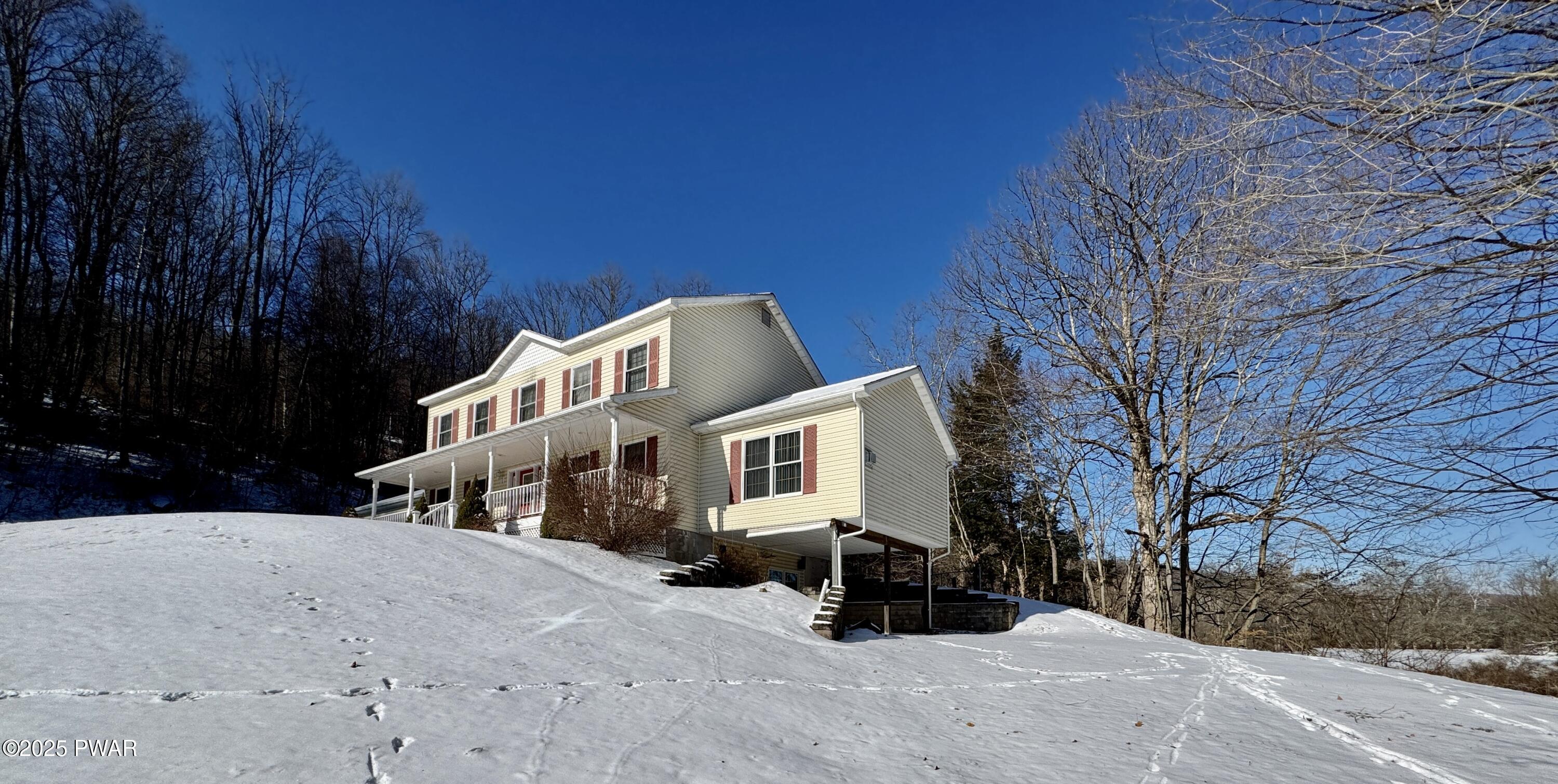 Two-story beige house with red shutters on a snowy hillside under a clear blue sky, surrounded by bare trees.