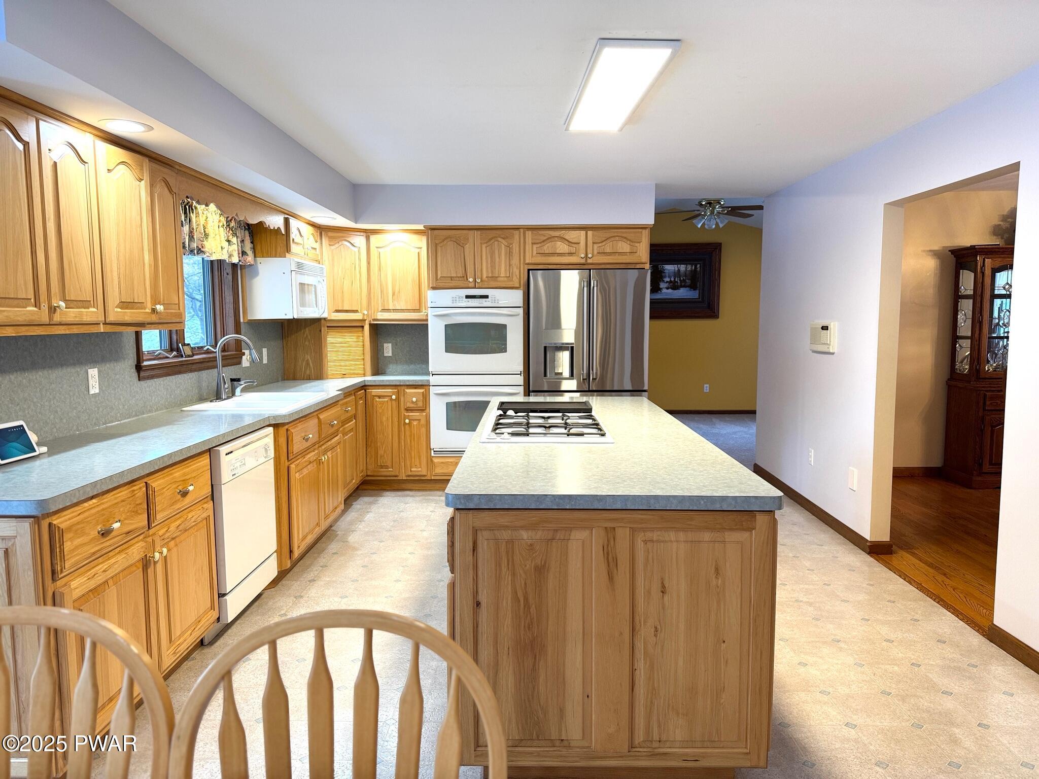 A bright kitchen with oak cabinets, an island cooktop, stainless steel refrigerator, built-in white oven and microwave, and a dining area opening to the side.