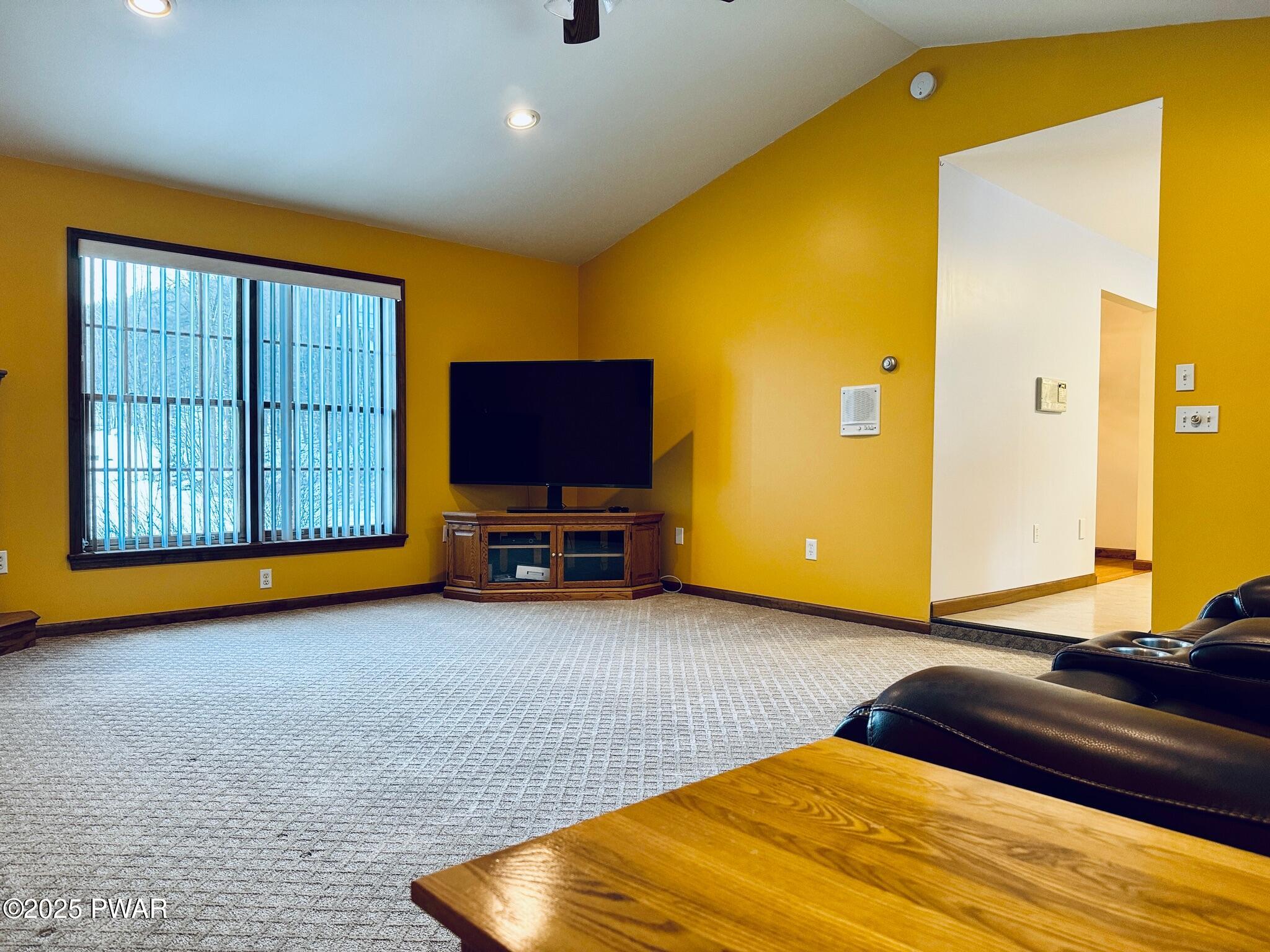 Spacious living room with yellow walls, carpeted floor, large window with vertical blinds, TV on a stand, and an open doorway to an adjacent area.