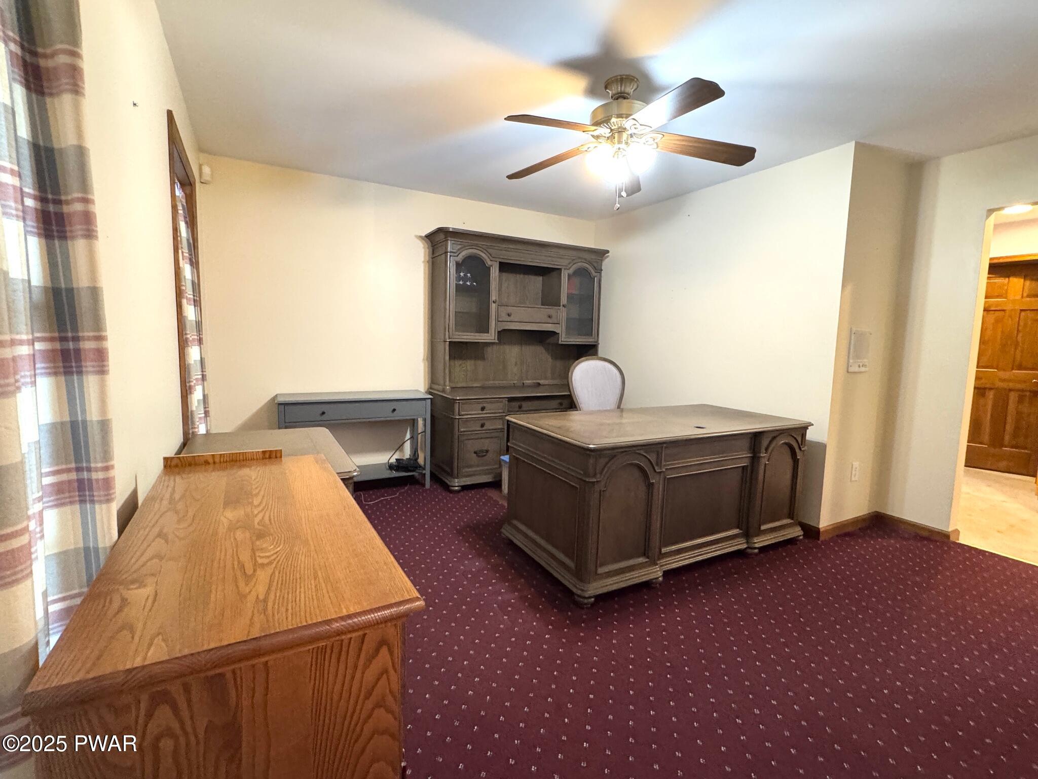 A furnished home office with a large wood desk, matching hutch, additional side table, ceiling fan, and patterned carpet.