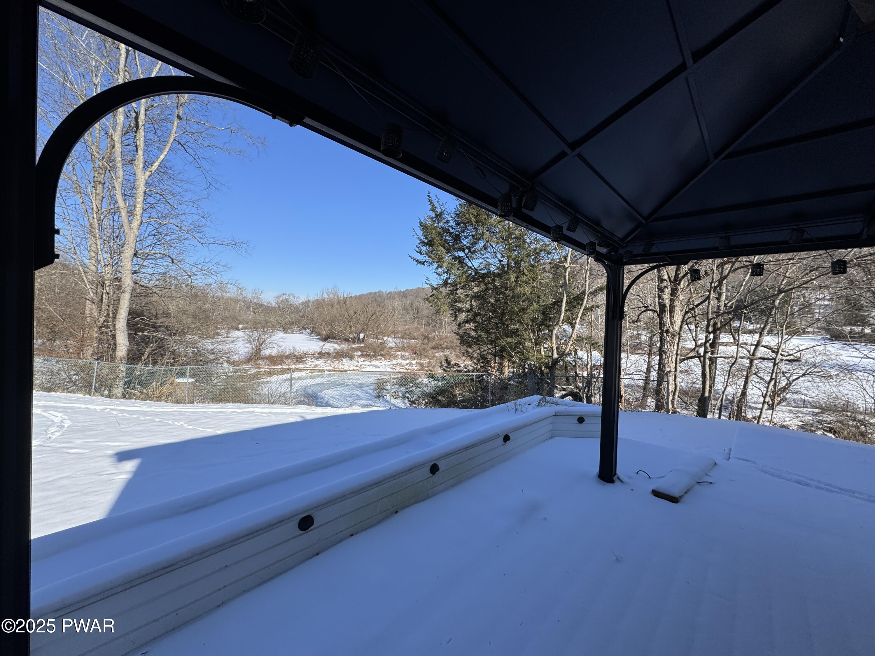 View from under a covered patio with a dark metal roof and posts, looking out over snow-covered ground, trees, and a winter landscape beyond a fence.