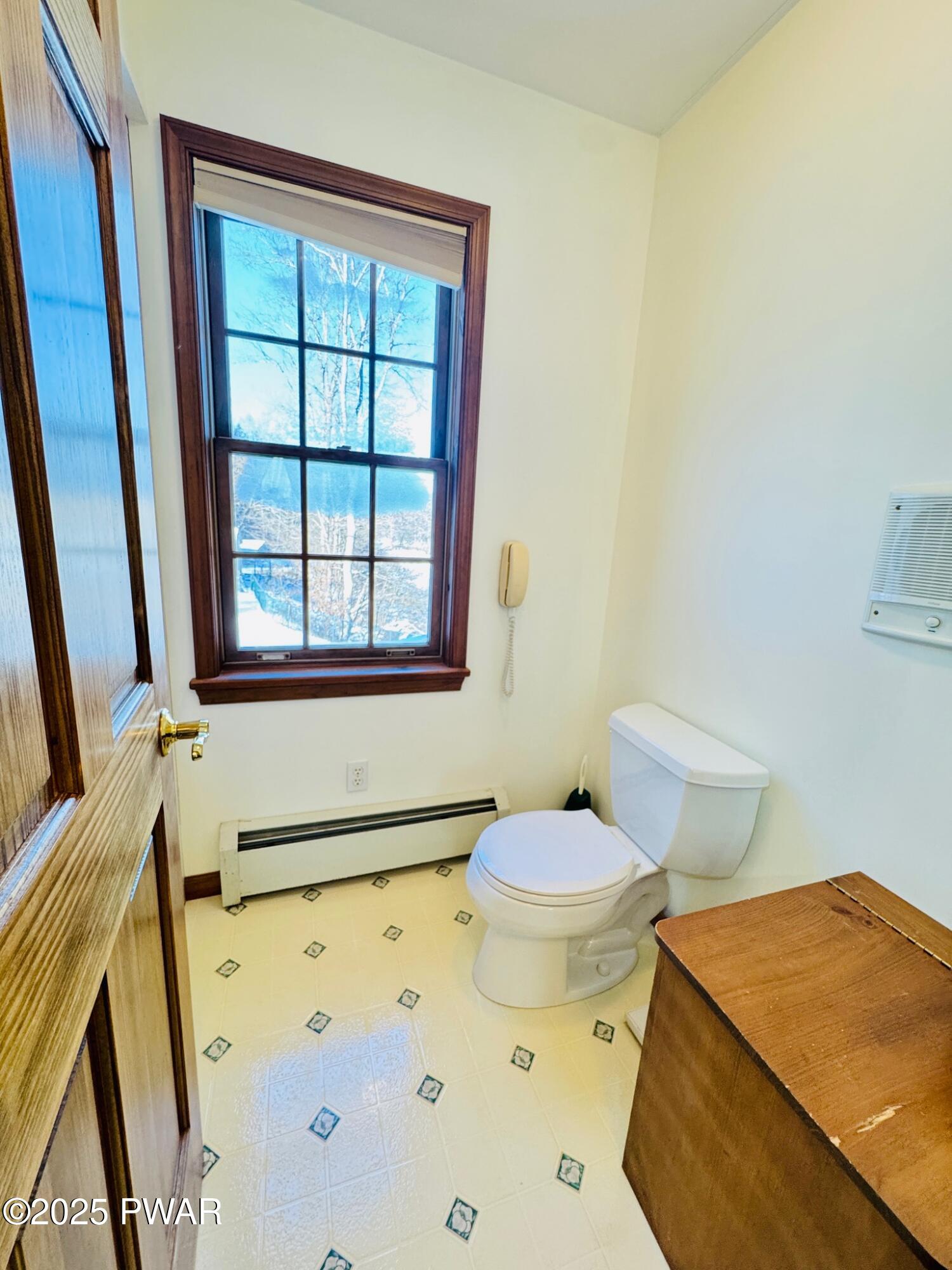 A small bathroom with a toilet beside a window, wood trim, tiled floor, and a wall-mounted heater.