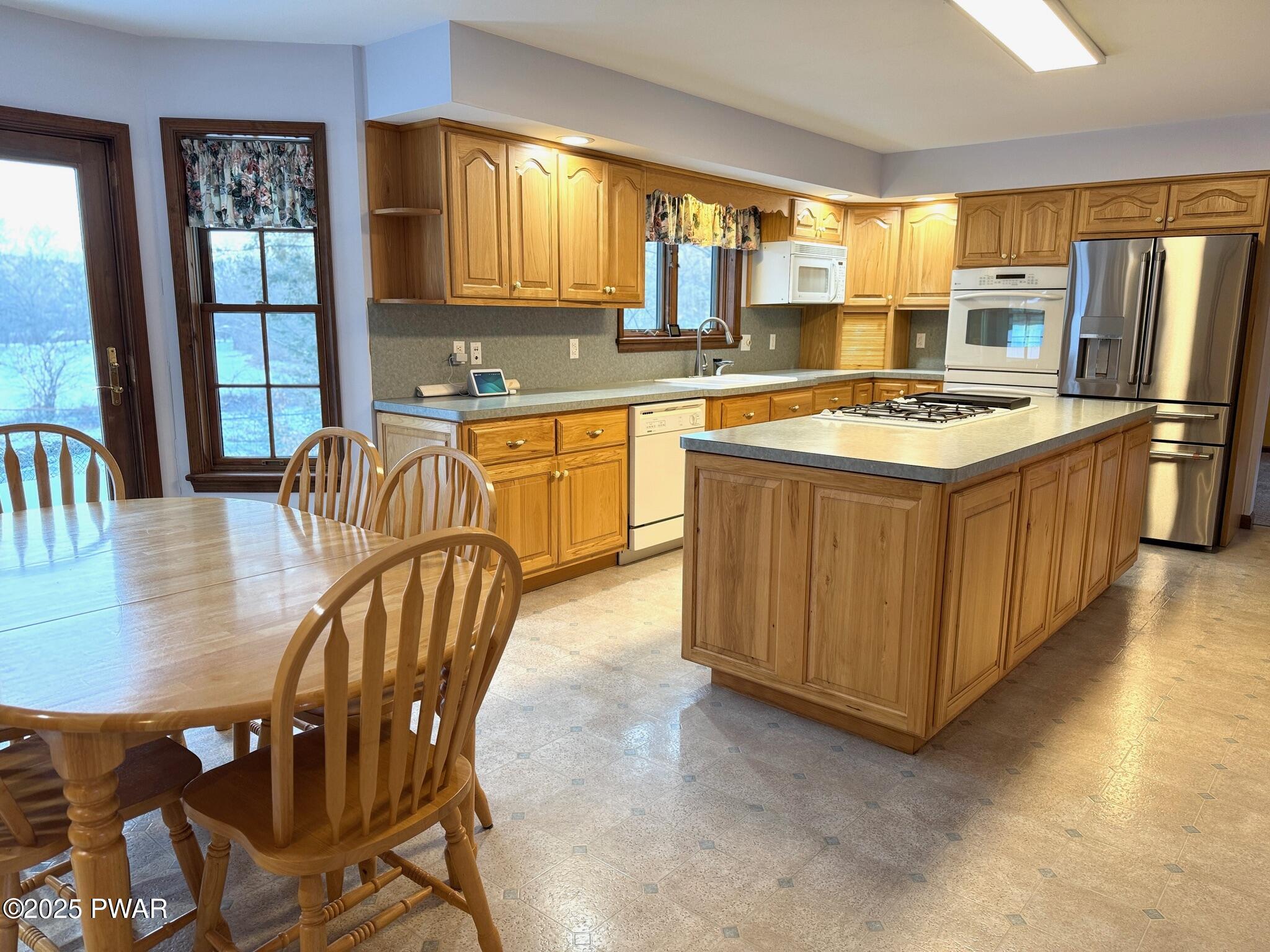 Spacious kitchen with oak cabinets, island, stainless steel refrigerator, and a wooden dining table by large windows and a glass door.