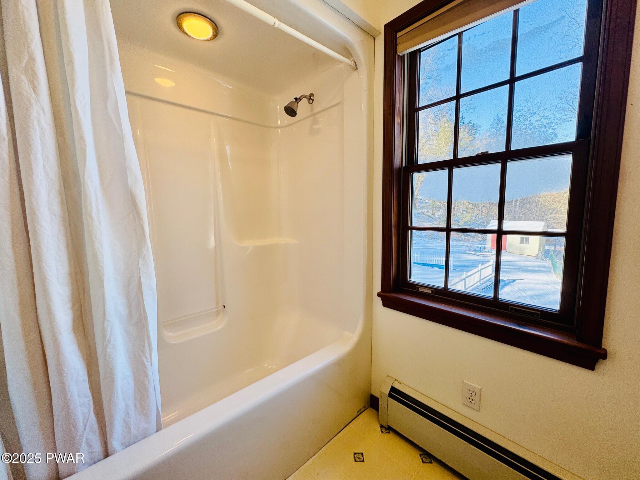 Bathroom with a white shower-tub surround and curtain beside a wood-trimmed window overlooking the outdoors.